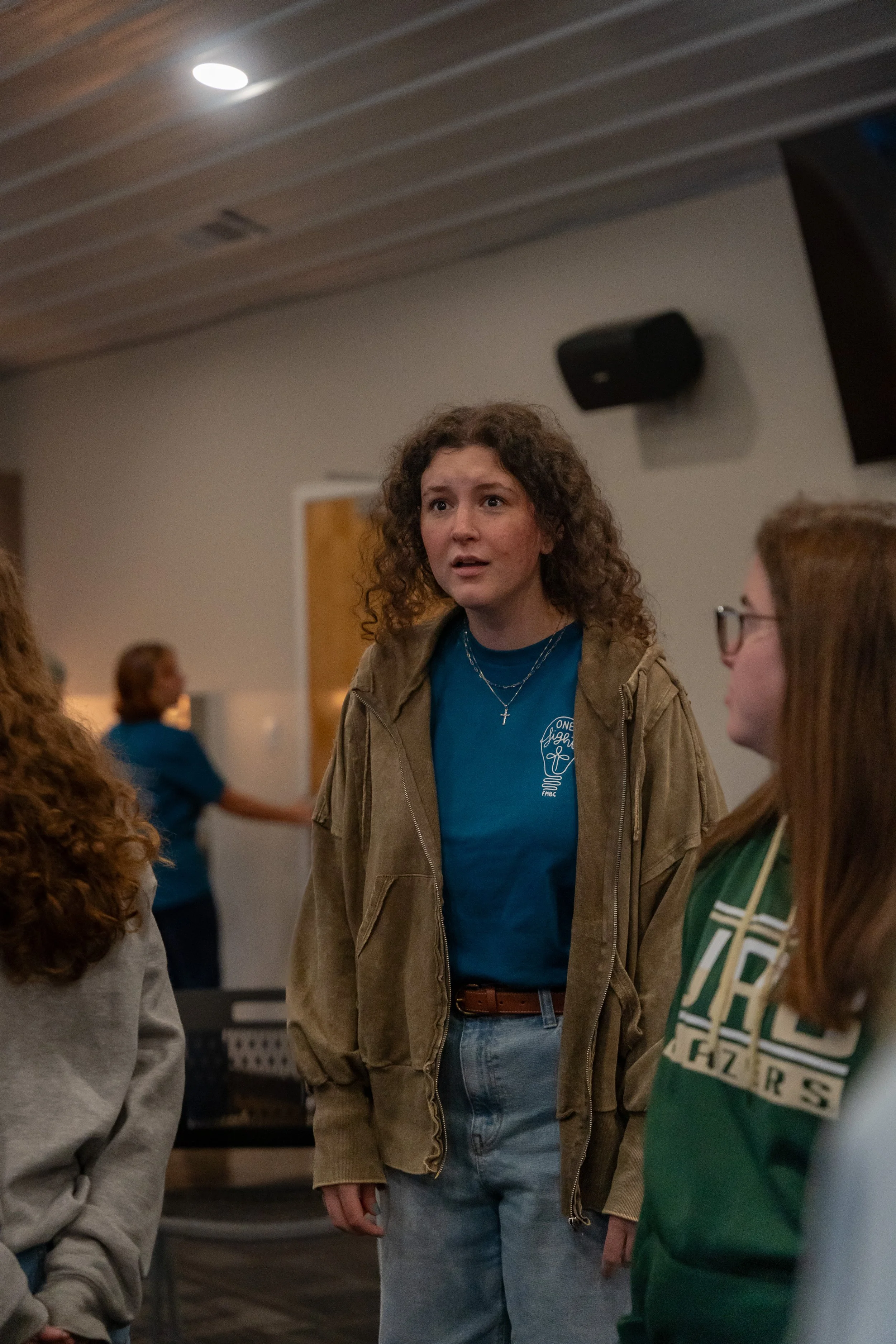A young woman with curly hair and a cross necklace talking to a group of people in an indoor setting, possibly a community center or church hall.