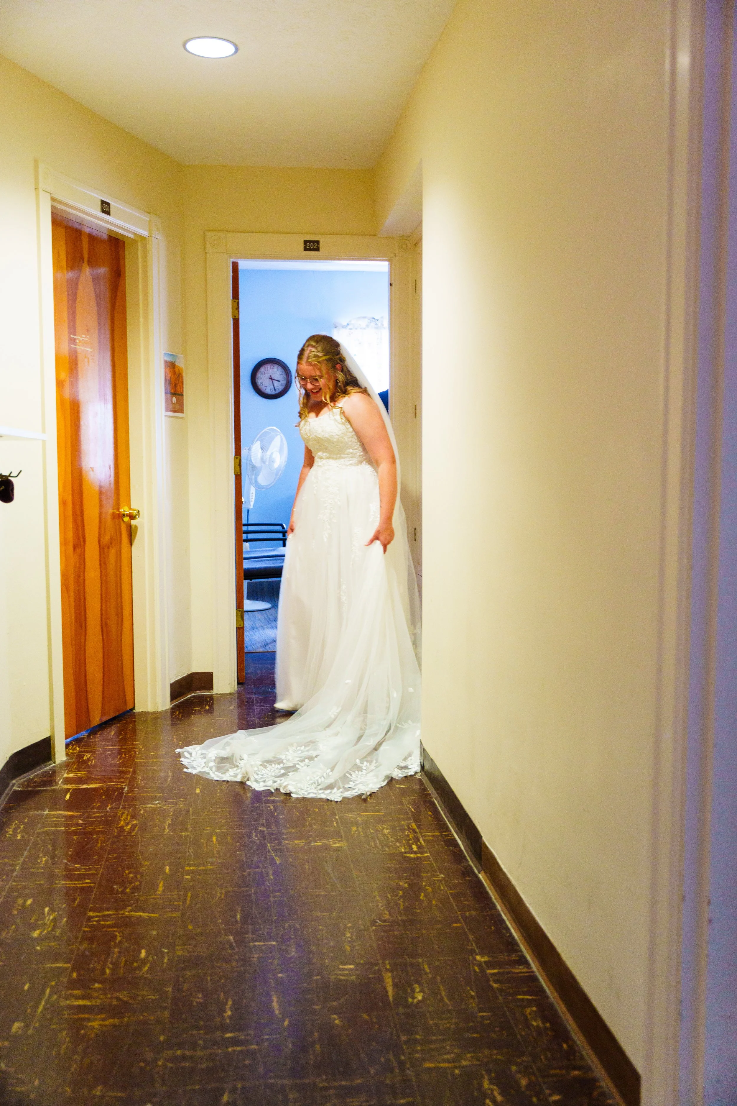 A woman in a wedding dress standing in a hallway, smiling and looking down, with a clock on the wall behind her and a fan visible in the background.
