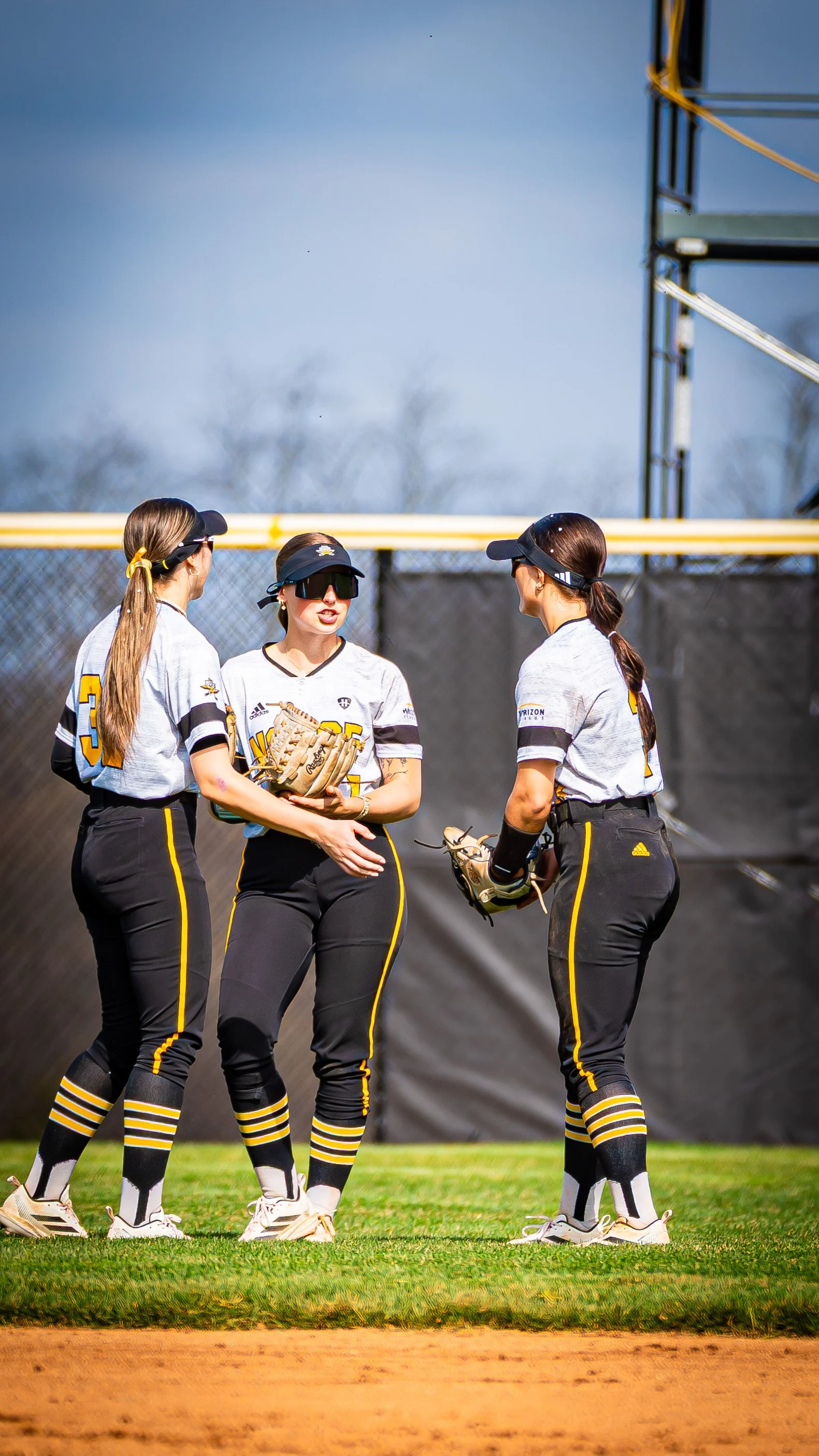Three female softball players in gray and black uniforms standing on the field and talking during a game.