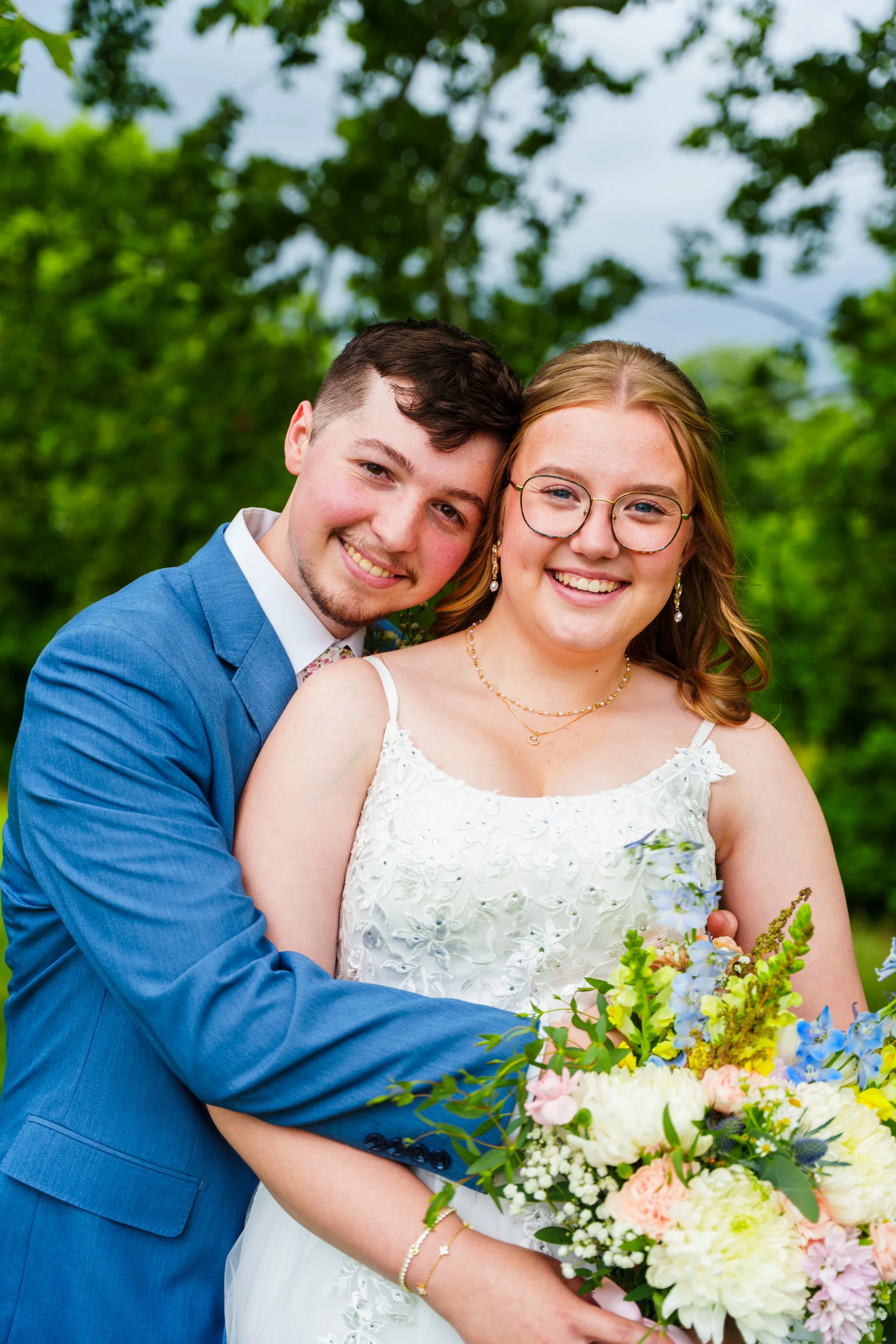 A happy couple in wedding attire embracing outdoors with lush green trees and sky in the background, holding a colorful bouquet of flowers.