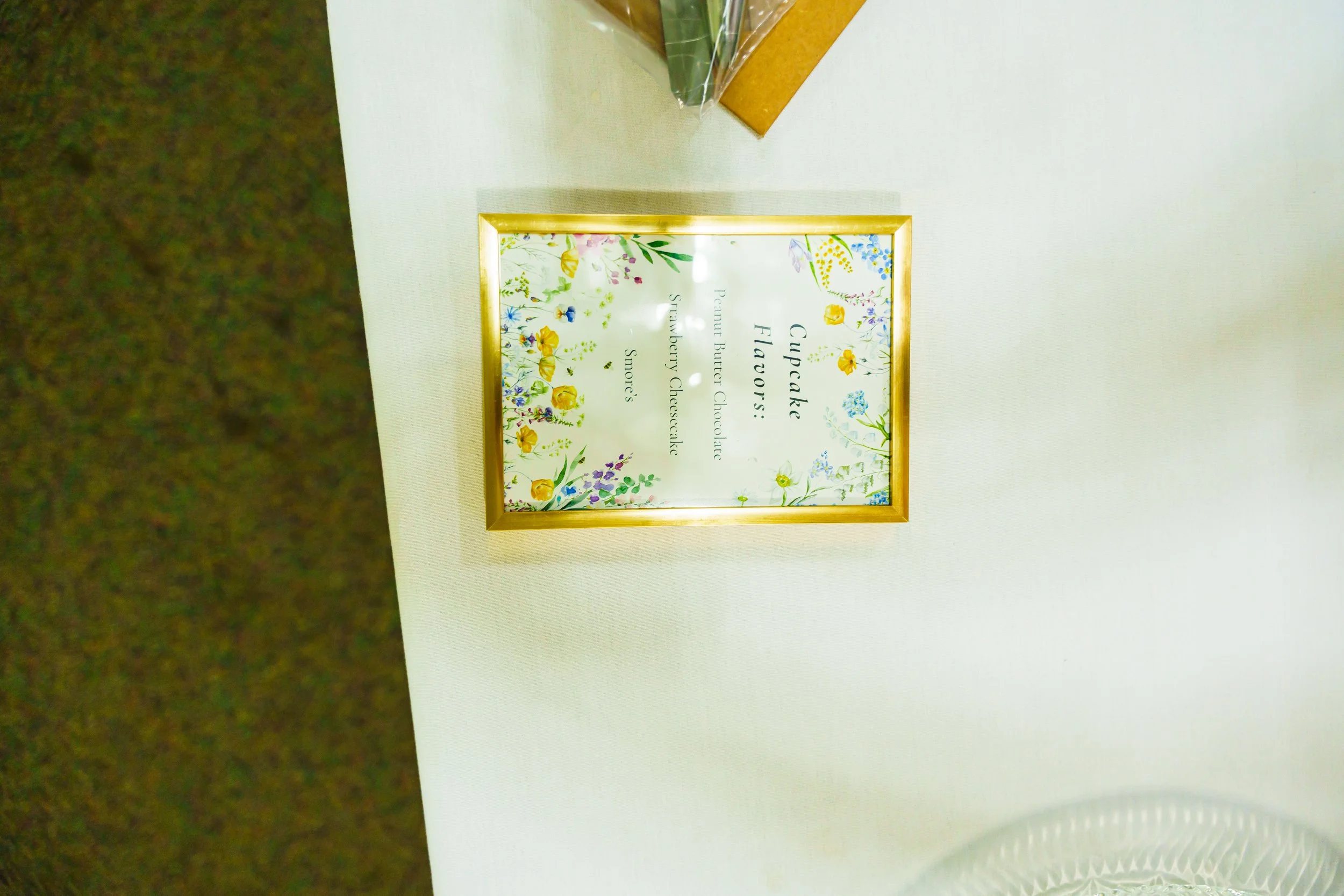 A framed floral sign on a white table. The sign has colorful flowers and green leaves, with text about cupcakes, cookies, and strawberry cheesecake.