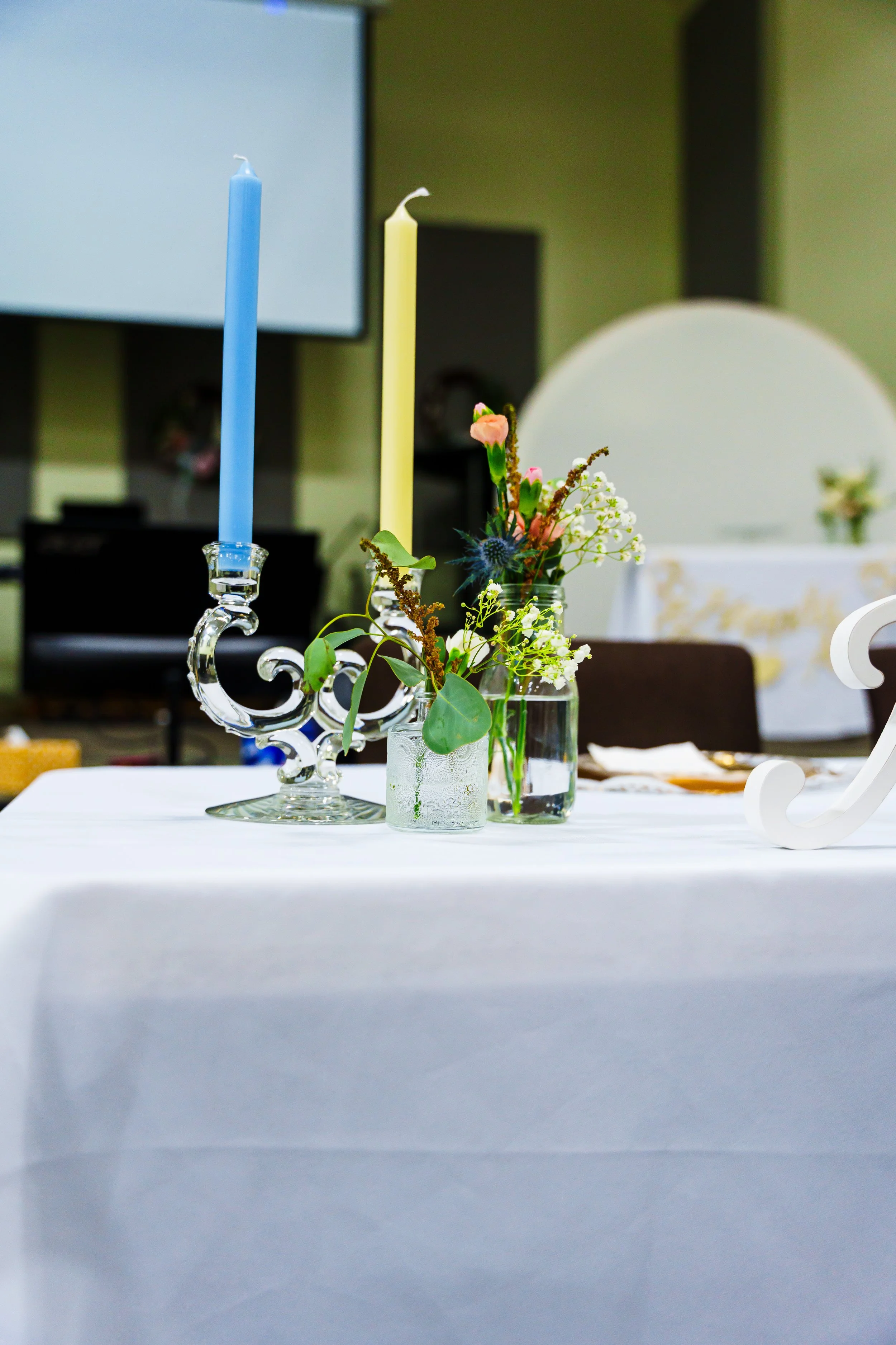 A white tablecloth-covered table with a glass vase holding a small flower arrangement, including pink and white flowers and greenery, and two tall candlesticks with blue and yellow candles, in a decorated event setting.