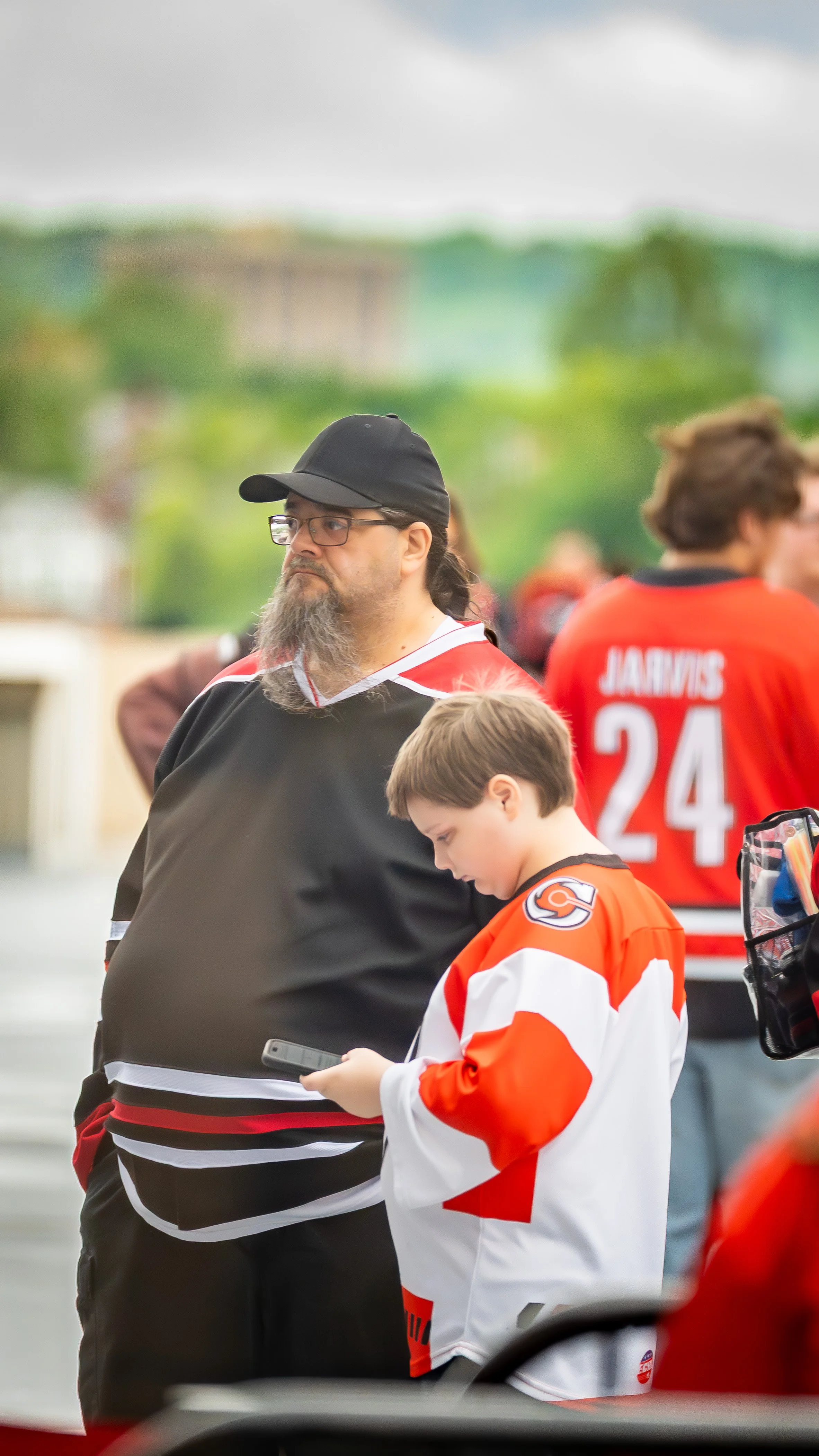 A man with glasses, a black cap, and a beard stands next to a young boy wearing an orange and black hockey jersey, both looking at a phone. In the background, other people in hockey jerseys are visible, with one displaying the name 'JACKINS' and the 