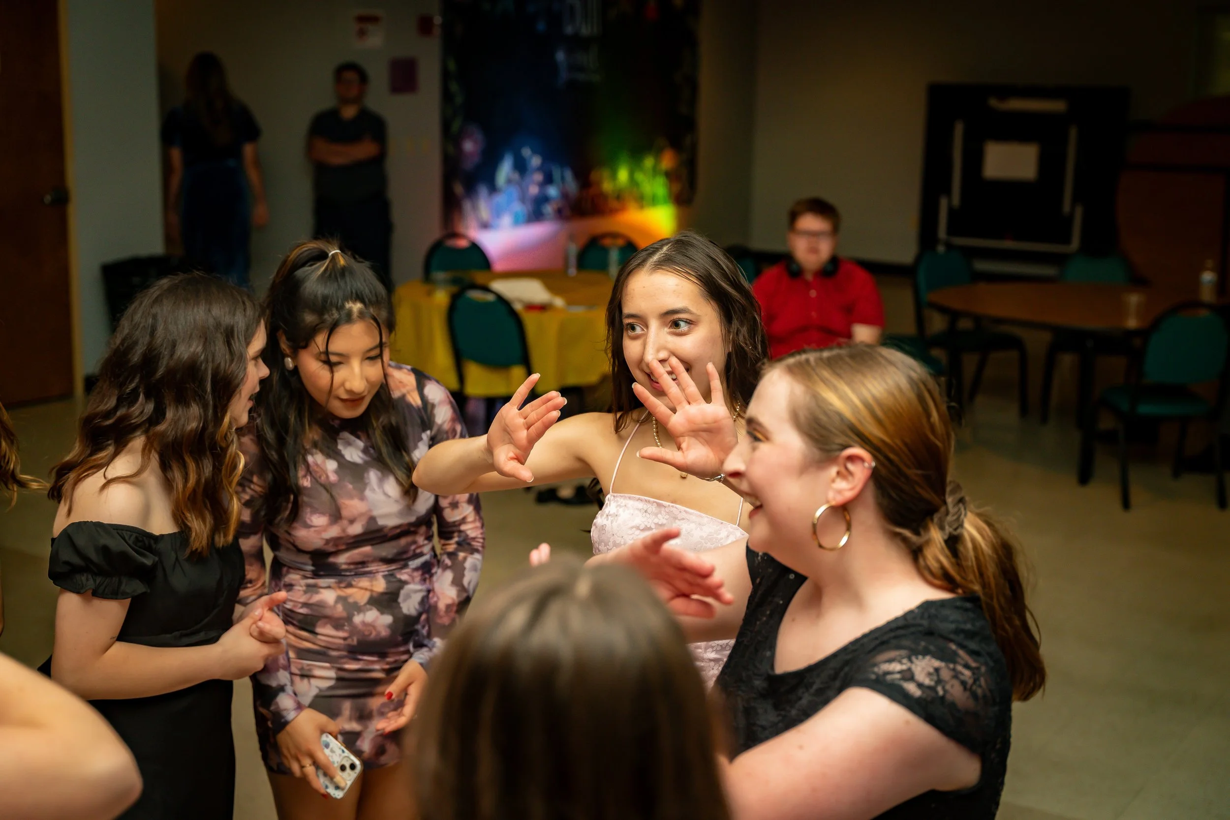 Group of young women socializing at a party or gathering in a room with tables and chairs, some women with braids, rings, and wearing stylish clothing, with a dark background and colorful lighting.