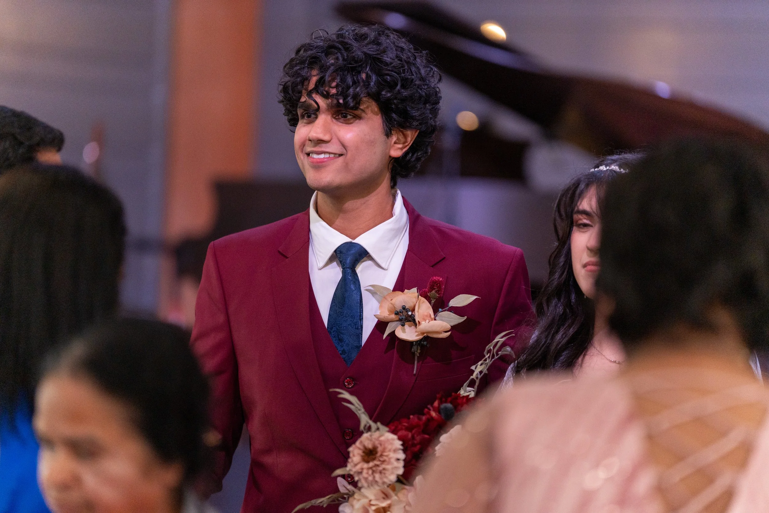 A man with curly hair wearing a maroon suit with a white shirt and blue tie, holding a flower boutonniere, smiling at a social gathering.