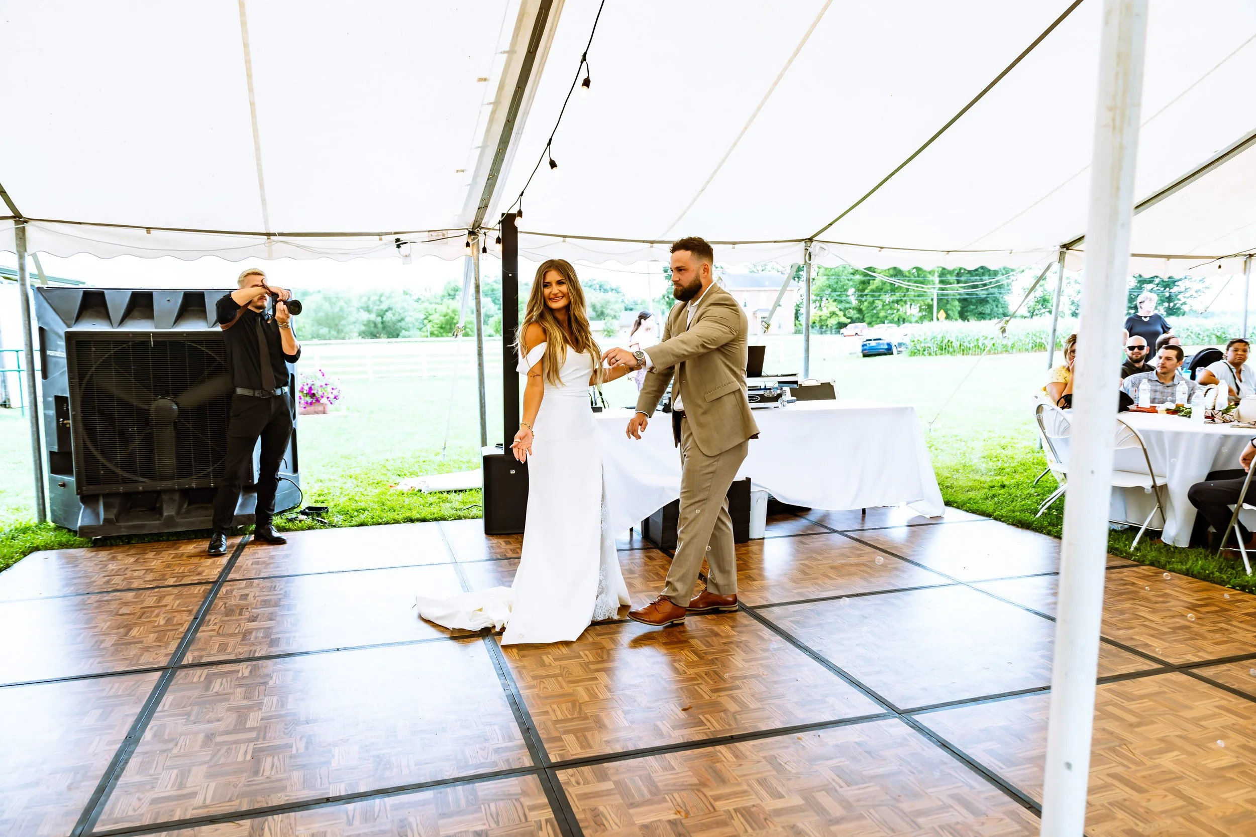 Bride and groom dancing at an outdoor wedding reception under a large white tent, with guests seated at tables and a photographer taking photos.