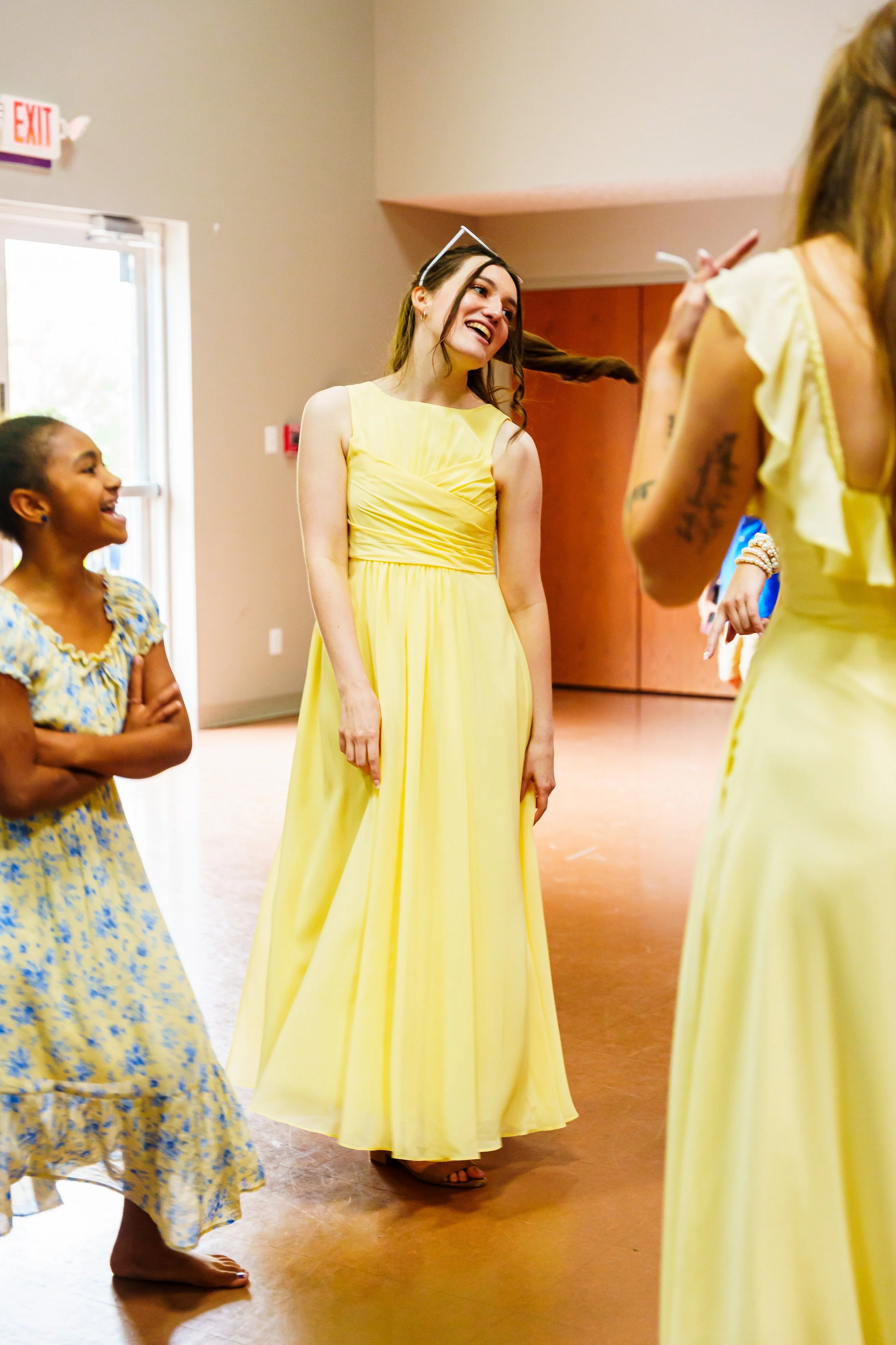 A group of women, mostly wearing yellow dress, standing indoors, some smiling and engaging in conversation.