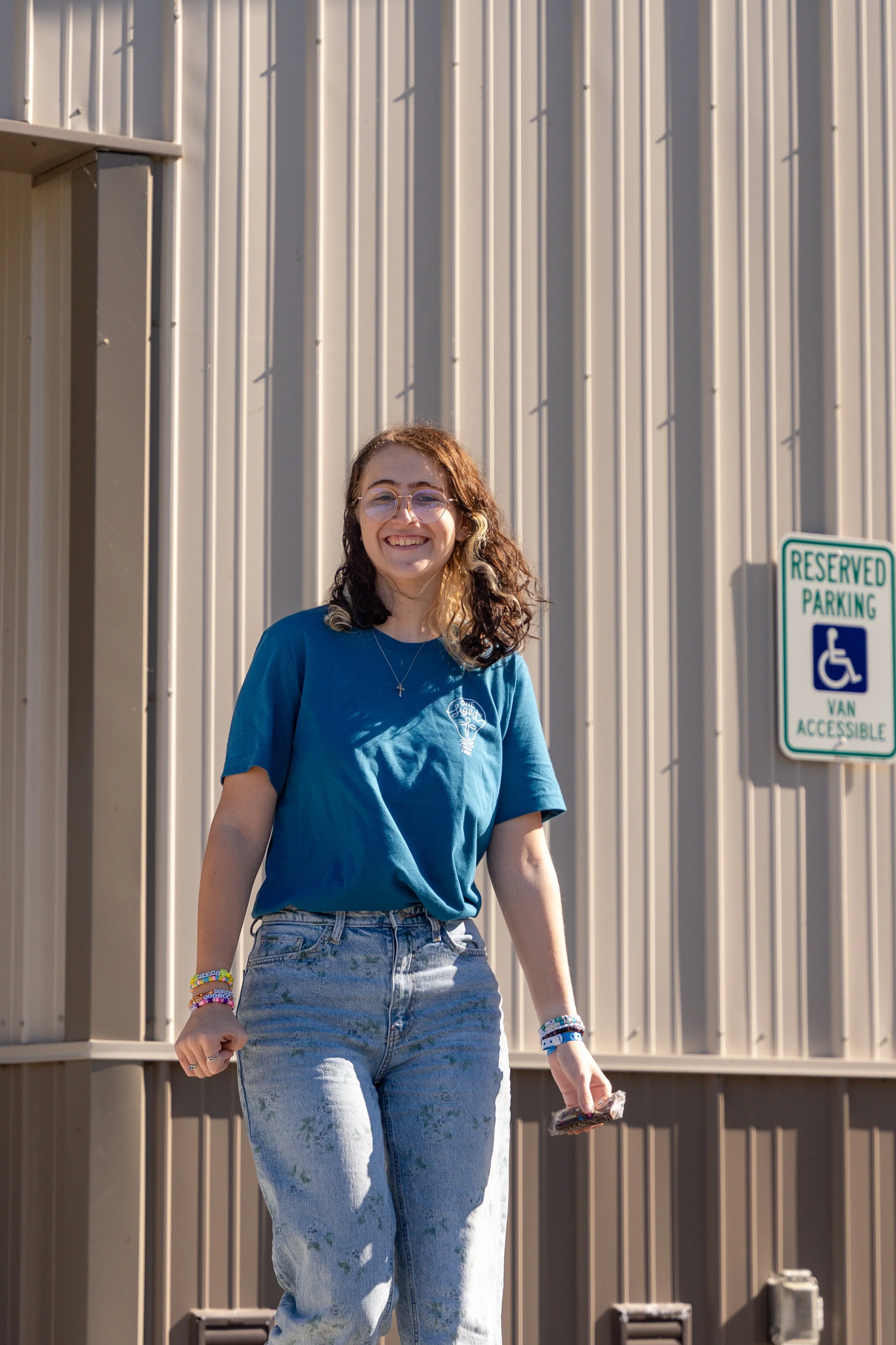 A young woman with curly hair wearing glasses, a blue t-shirt, and light blue jeans, smiling while walking outdoors near a beige wall with vertical lines. She has colorful bracelets on her left wrist and is holding a small object in her right hand.