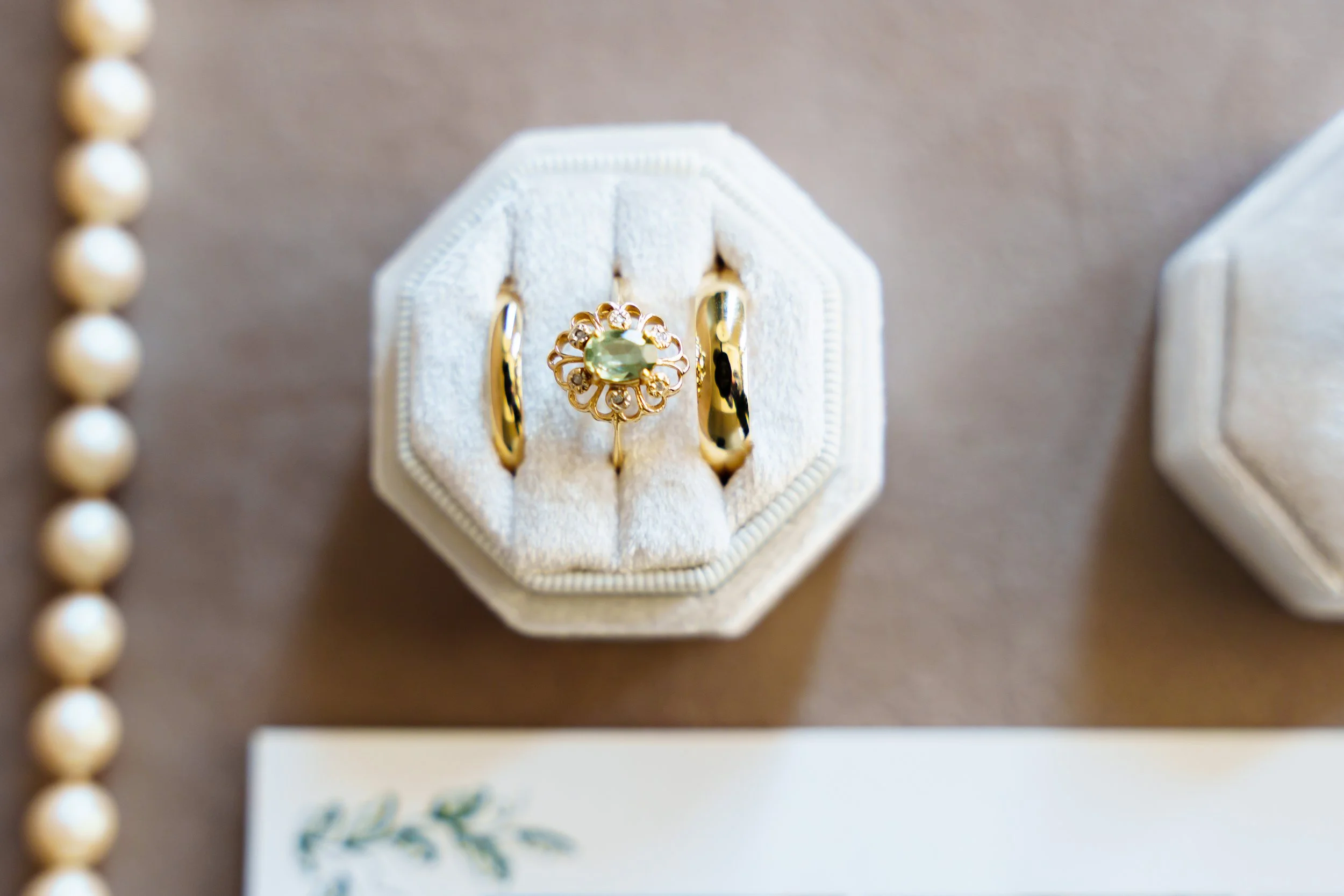 A close-up of a jewelry ring with a yellow gemstone and small diamonds on a white velvet ring box, surrounded by a pearl necklace and a blurred card with a floral design.