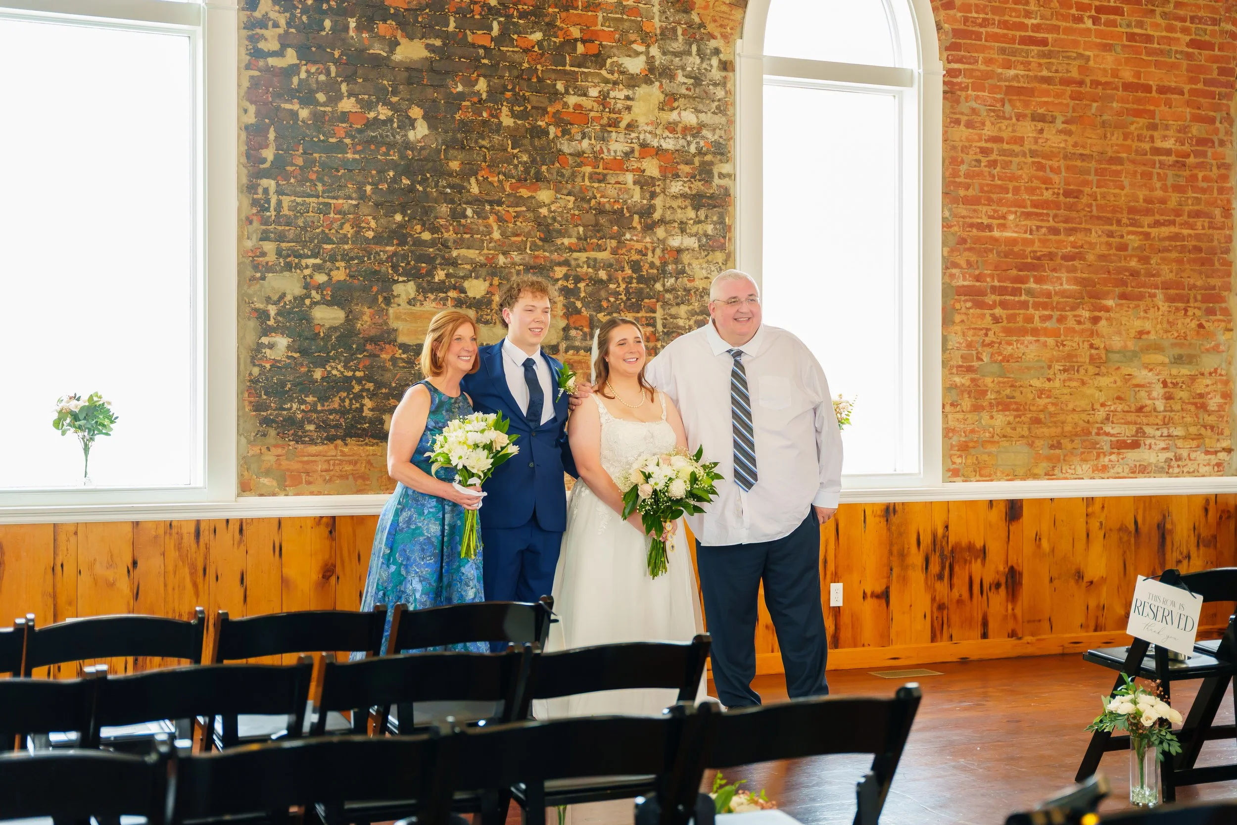 A bride and groom with two adults, all smiling, standing together at a wedding ceremony inside a room with exposed brick walls and large windows. The bride is holding a bouquet, and the groom is wearing a blue suit. The room has black chairs and a re