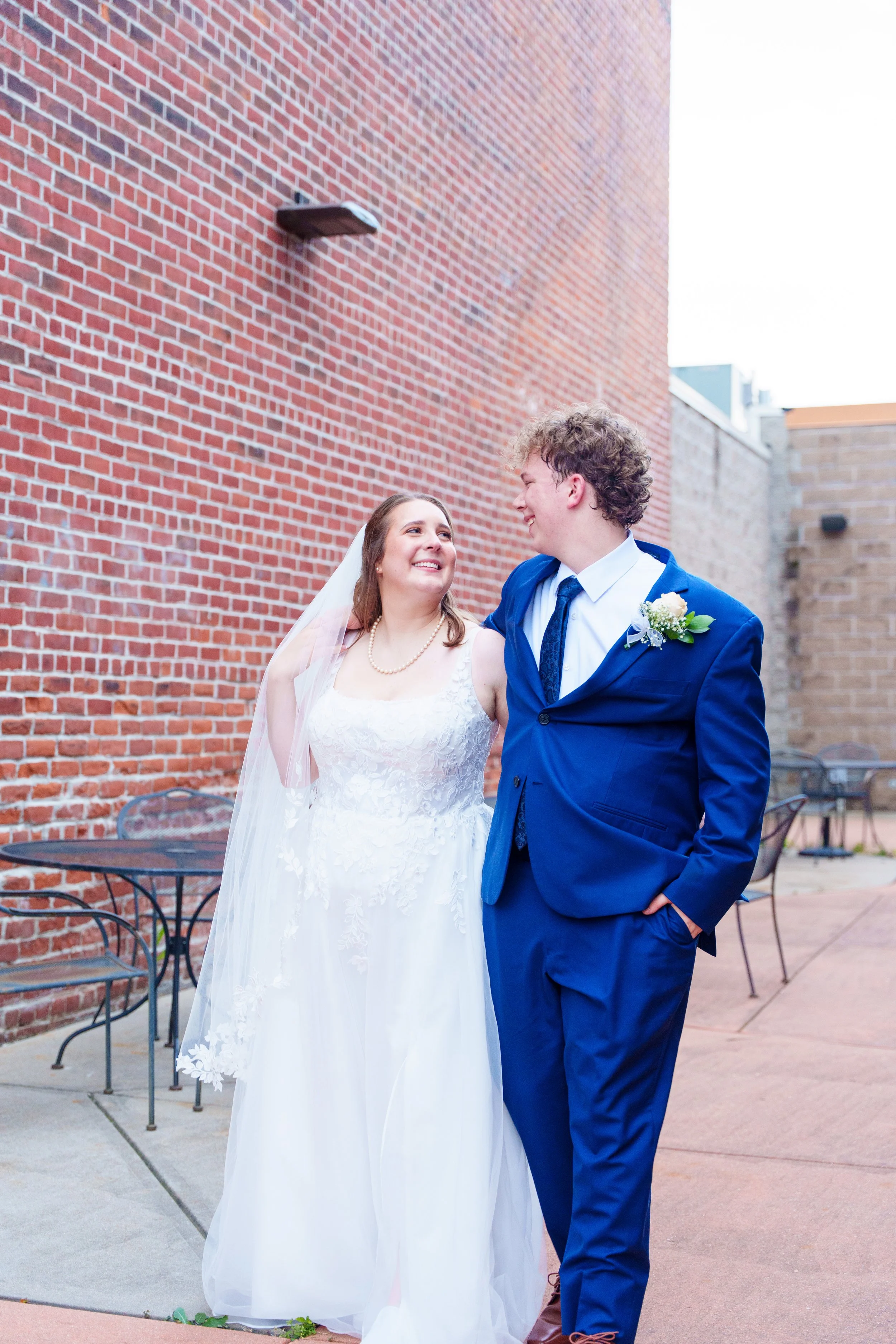 A bride and groom smiling at each other outdoors near a red brick wall, dressed in wedding attire. The bride is in a white gown with a veil and pearl necklace, and the groom is in a blue suit with a white shirt and blue tie, with a boutonniere on his