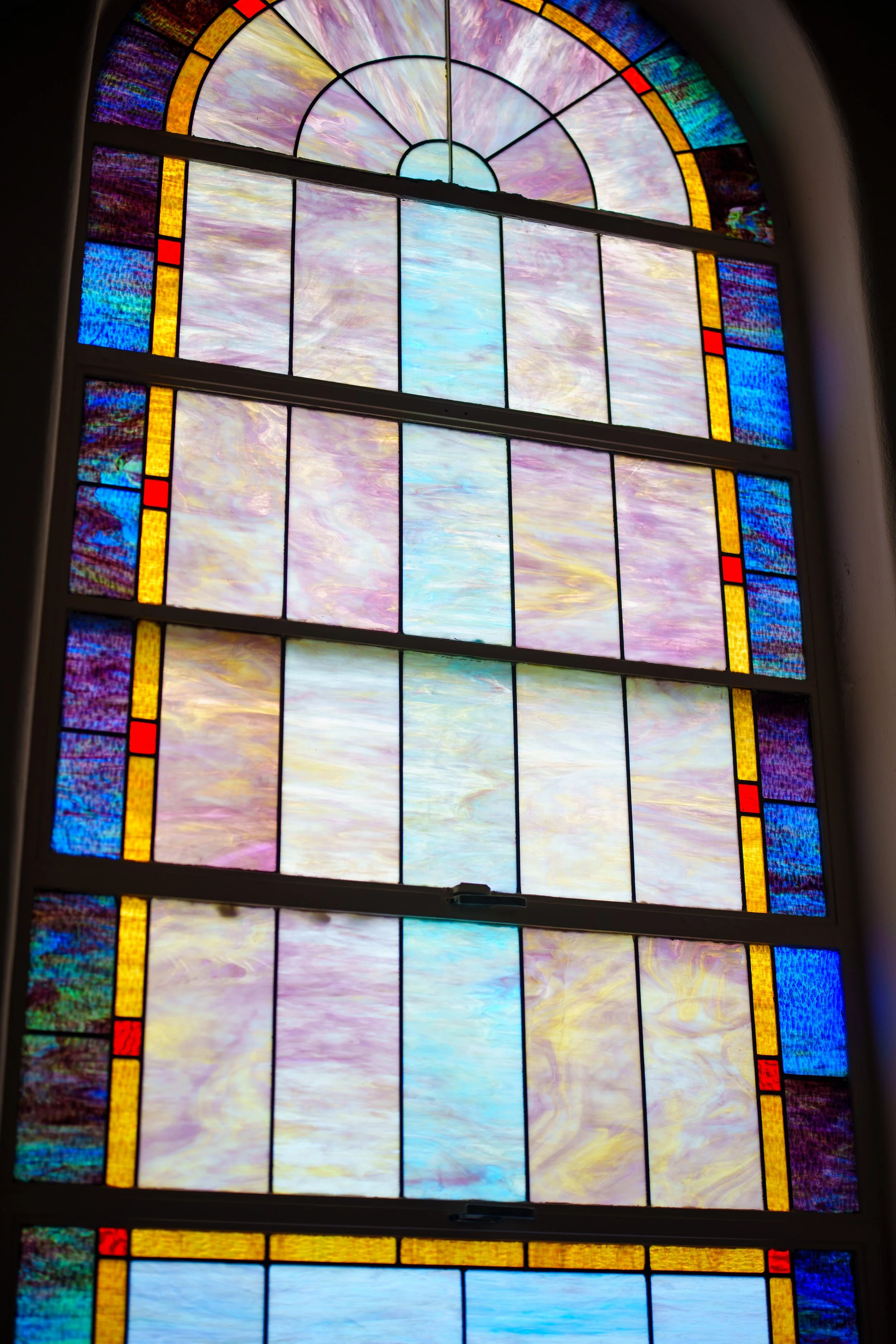 Colorful stained glass window with pastel clouds and abstract patterns in purple, pink, blue, and yellow hues, framed with red, yellow, and blue border segments.