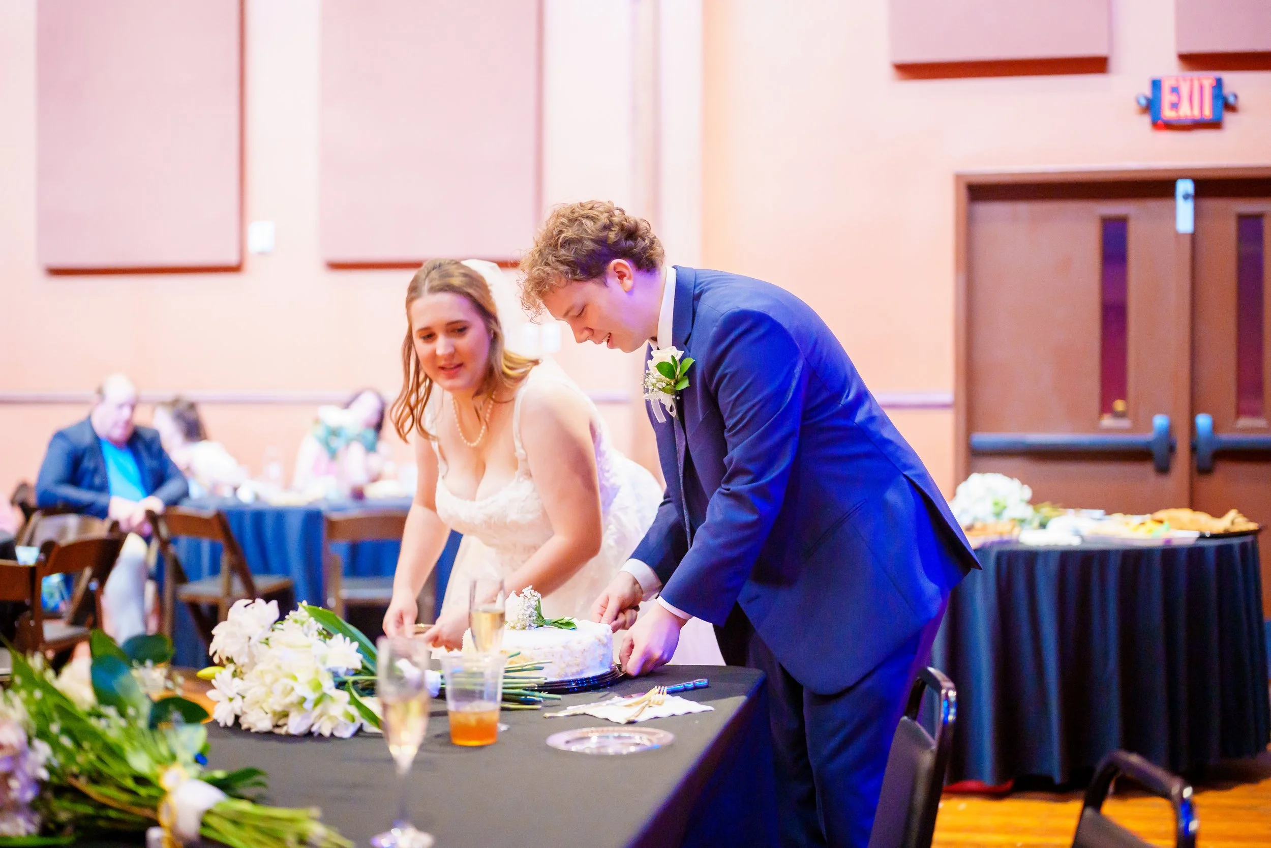 A bride and groom cut a wedding cake at their reception. The bride wears a white wedding dress and the groom is in a blue suit.