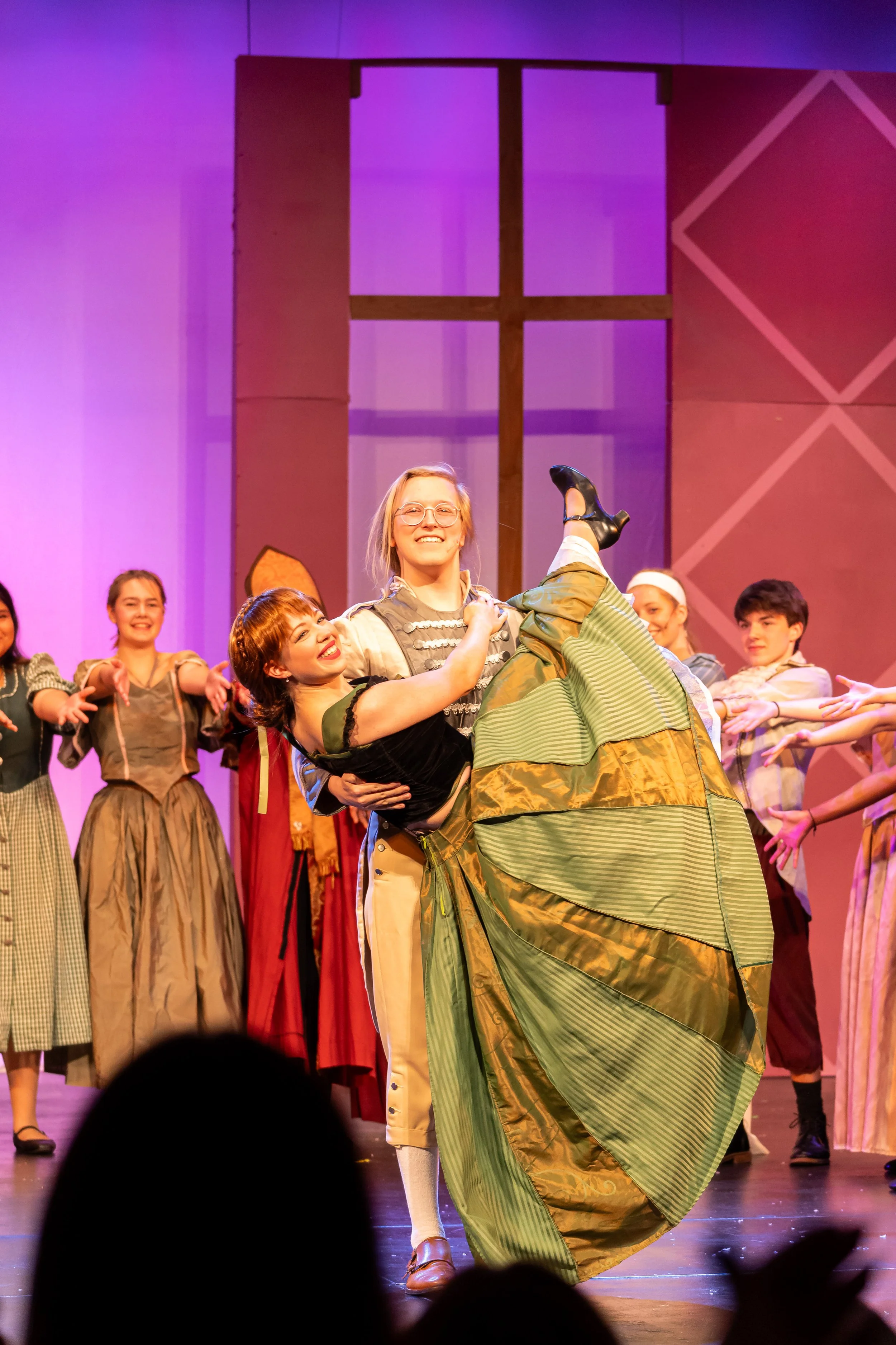 Stage performance featuring actors in historical costumes, with one actor in a green dress and others smiling in the background.