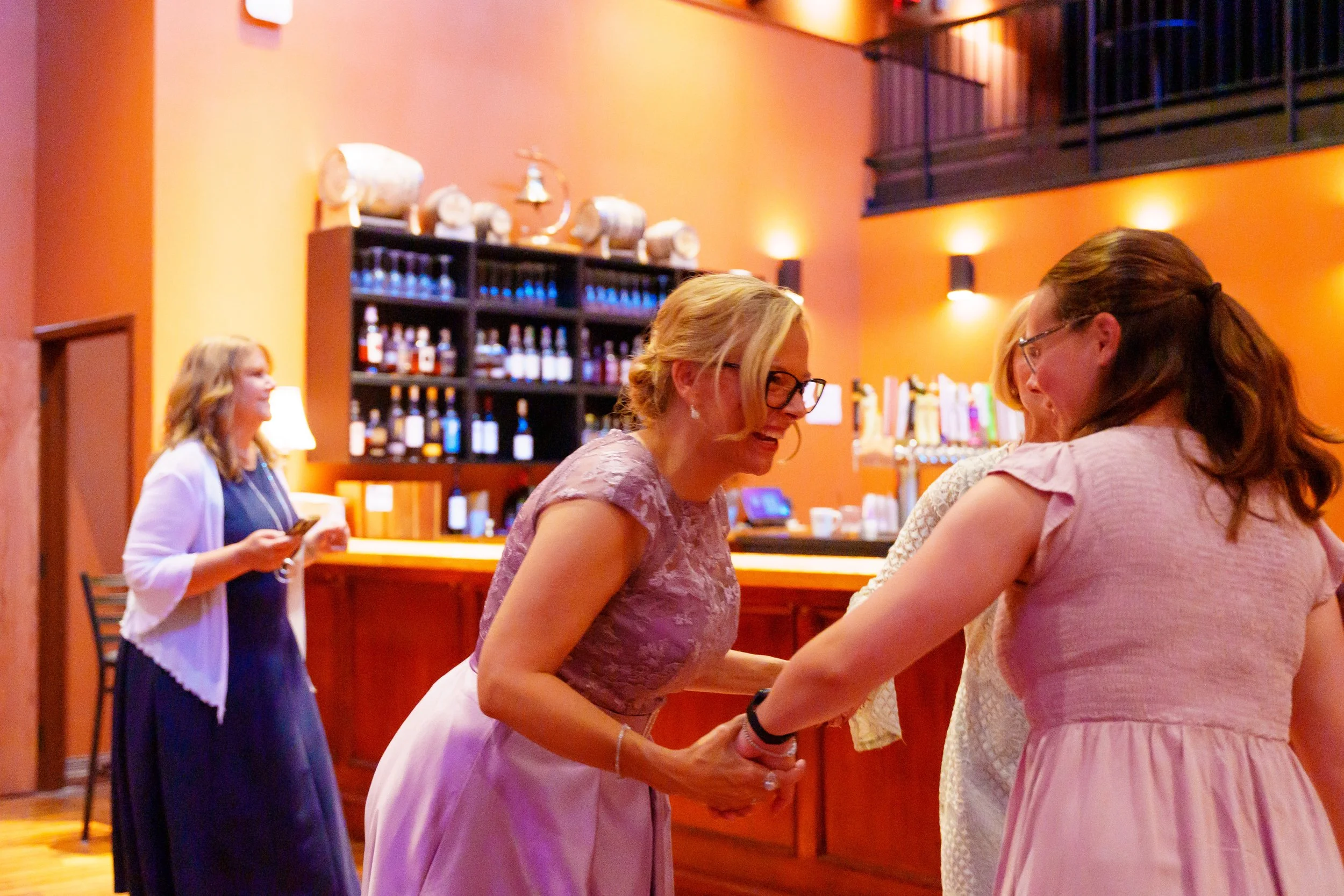 A group of women at a social gathering in a warmly lit bar or restaurant, shaking hands and smiling.