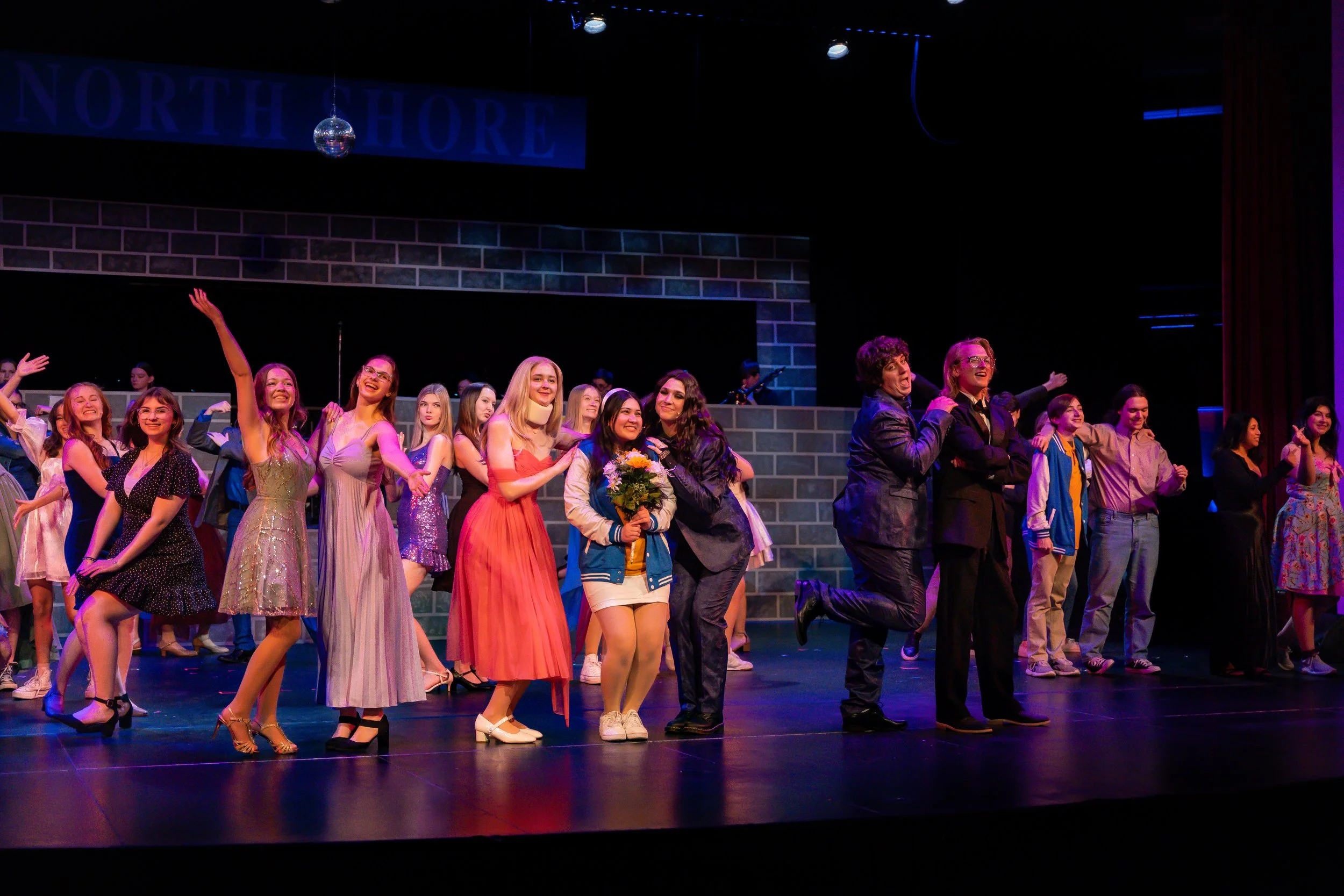 Group of performers on stage at a theater, with a sign reading 'North Shore' above them. Some performers are dressed in vintage-style dresses, and one woman holds a bouquet of flowers. The scene appears to be the end of a performance or curtain call.