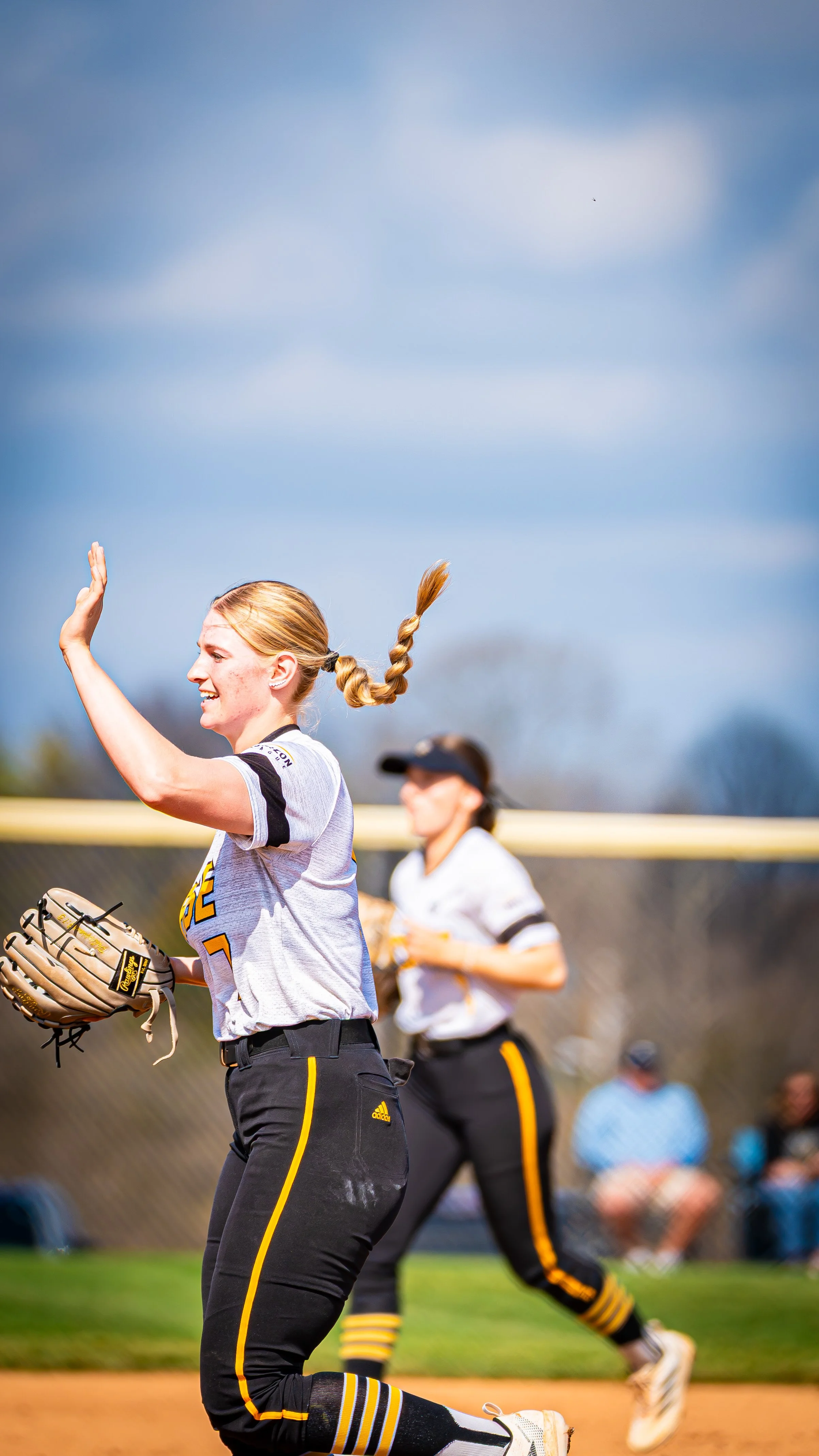 A young female softball player with blonde hair in a braid, wearing a uniform, raising her hand during a game on a sunny day with a blue sky.