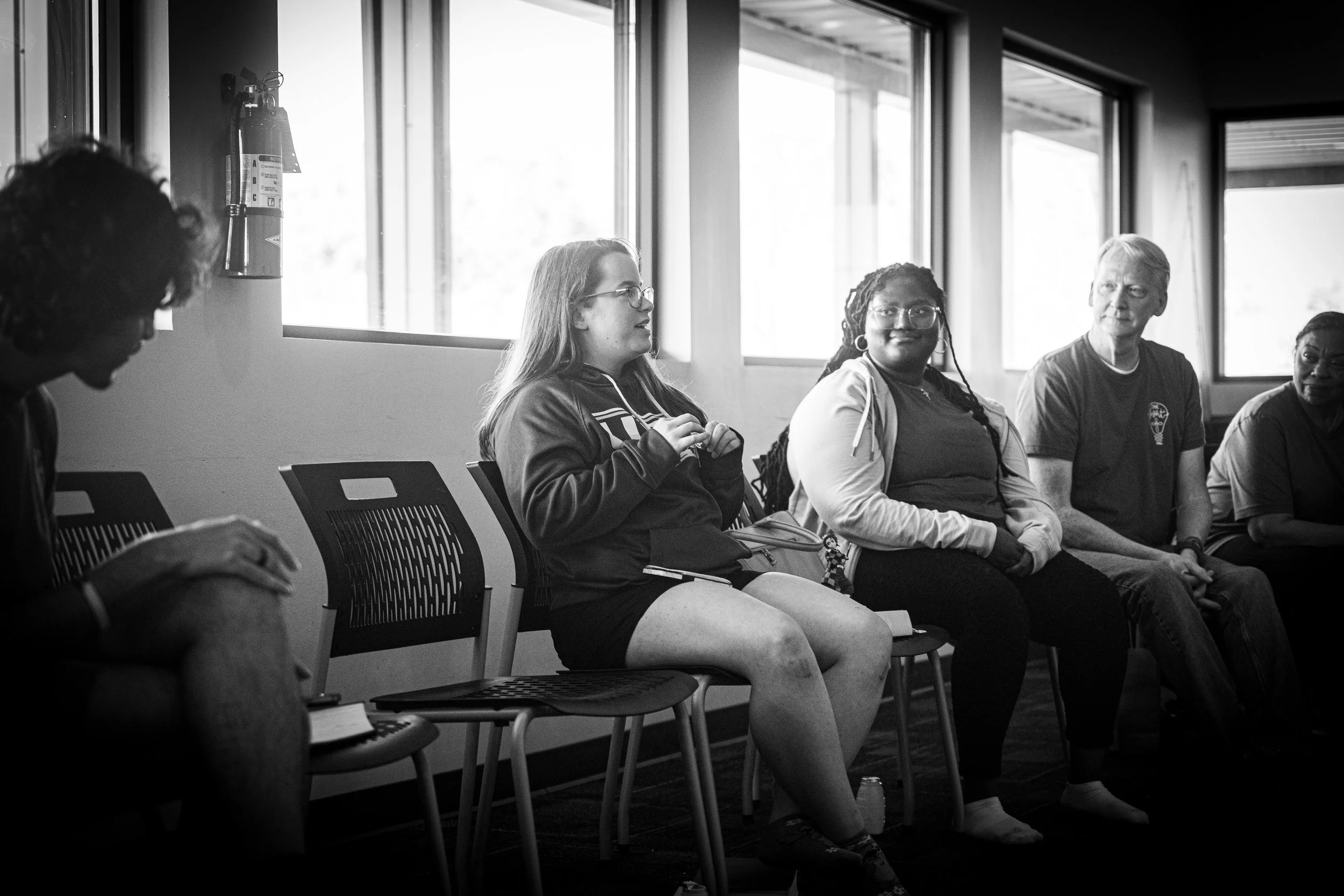 Group of five diverse people sitting on chairs in a room with large windows, engaged in a conversation or listening.