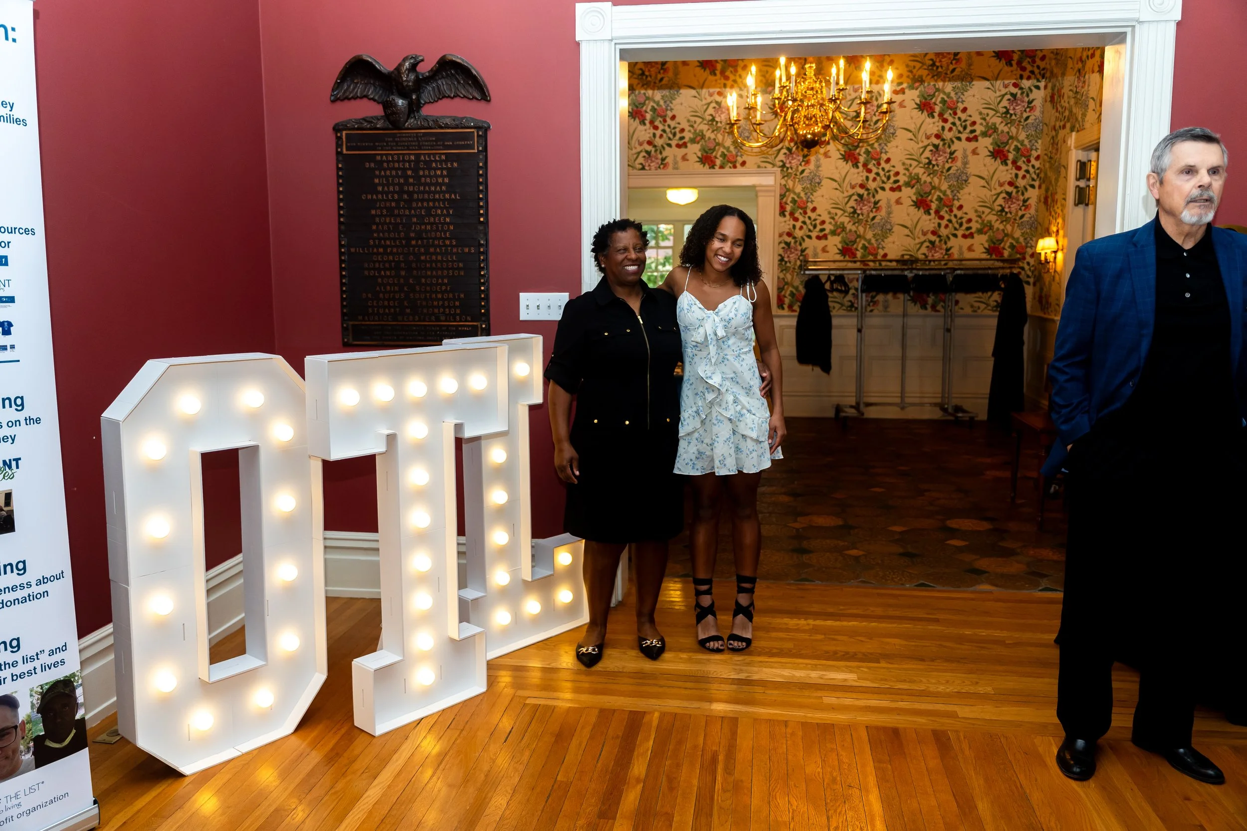 Two women standing next to illuminated letters spelling 'LOVE' at an indoor event, with one woman wearing a black dress and the other in a floral dress, both smiling.