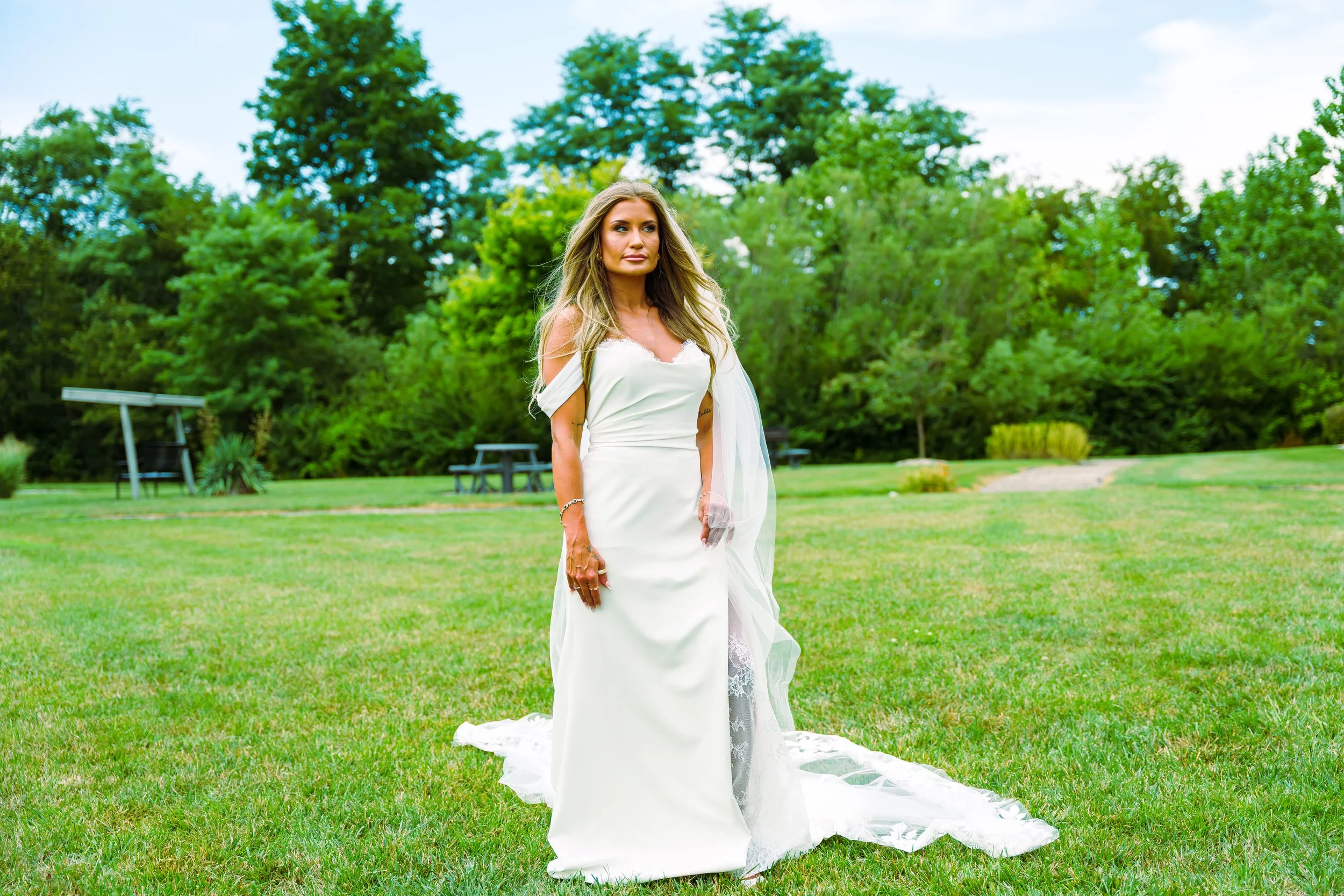 A blonde woman in a white wedding dress standing on a grassy field with trees in the background.