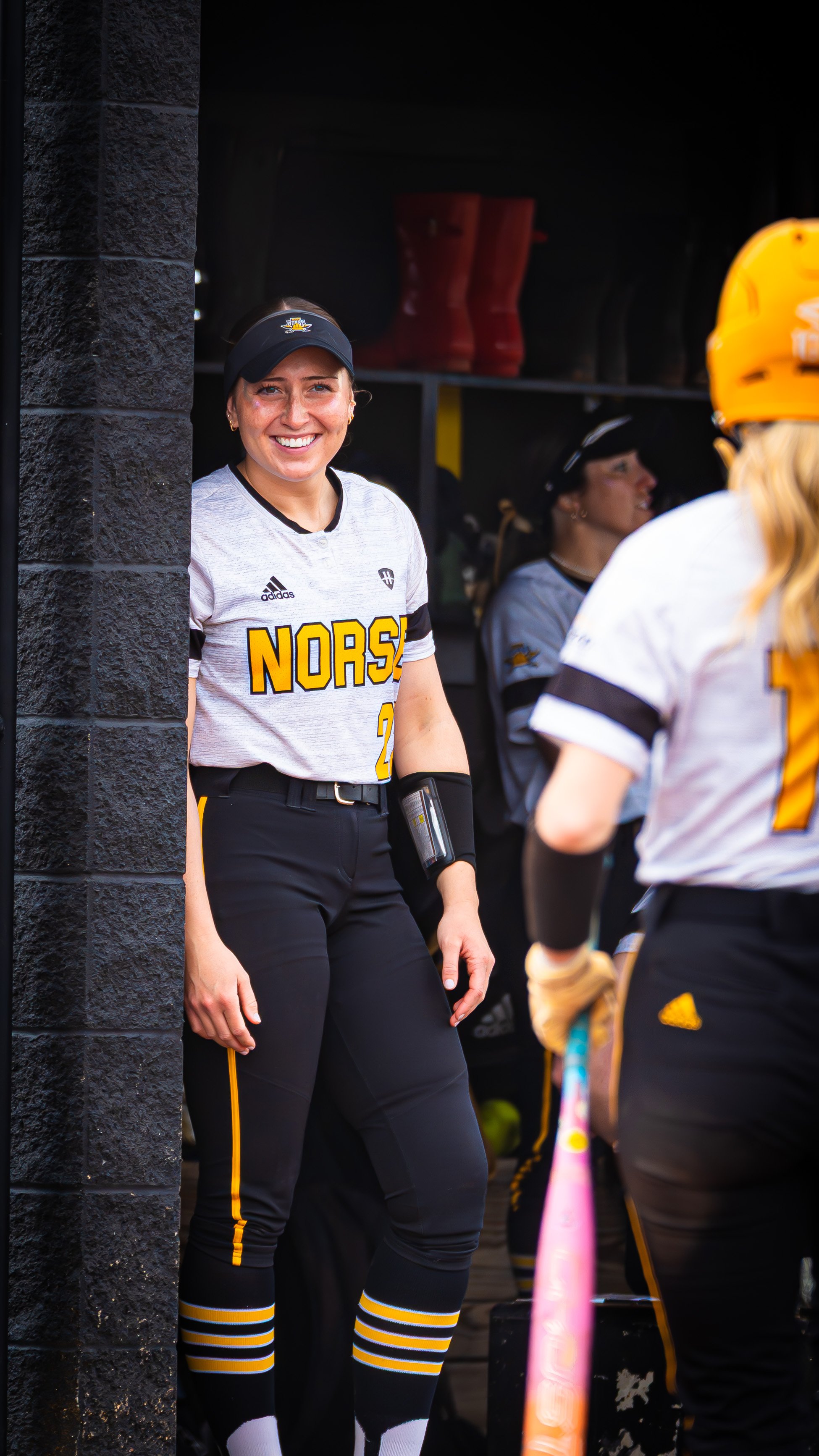 Female softball player smiling while standing in a dugout with teammates behind her, wearing a gray and black uniform with 