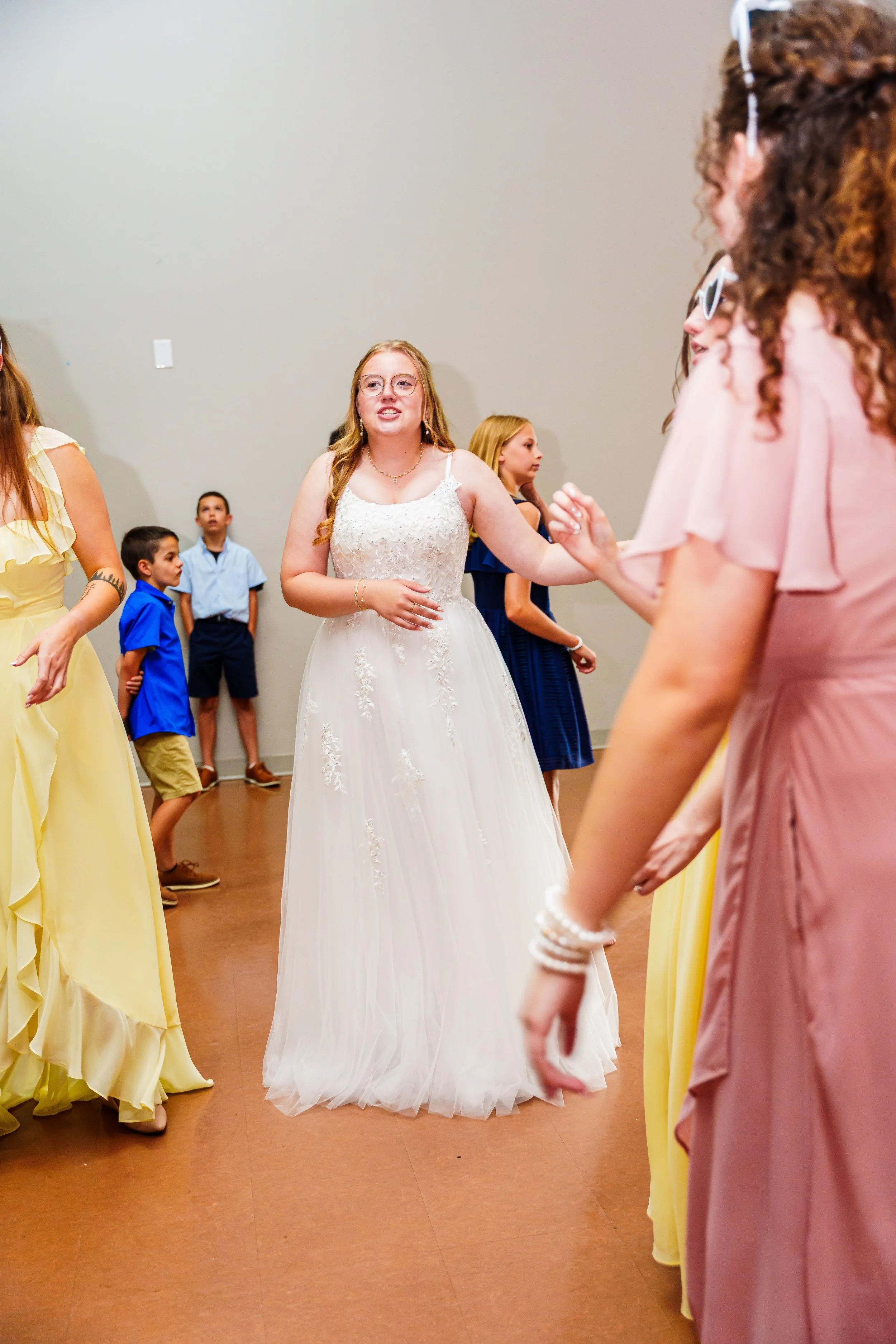 A bride in a white wedding dress dancing with a group of women at a wedding reception, surrounded by children in formal attire.