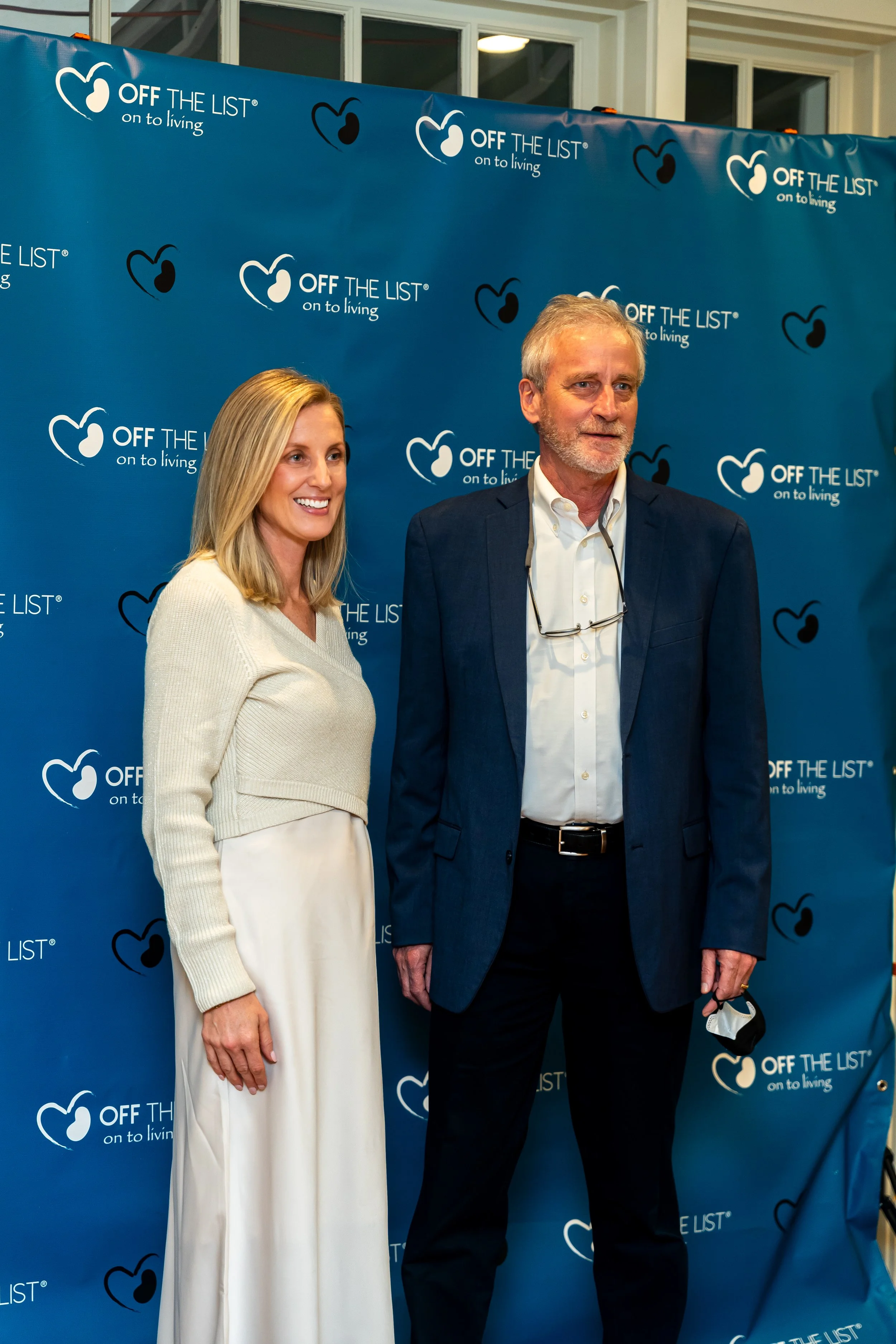 Two people, a woman and a man, standing in front of a blue backdrop with white and black heart-shaped logos and the text 'OFF THE LIST on to living.' The woman has blonde hair and is wearing a cream-colored sweater and white pants. The man has gray h
