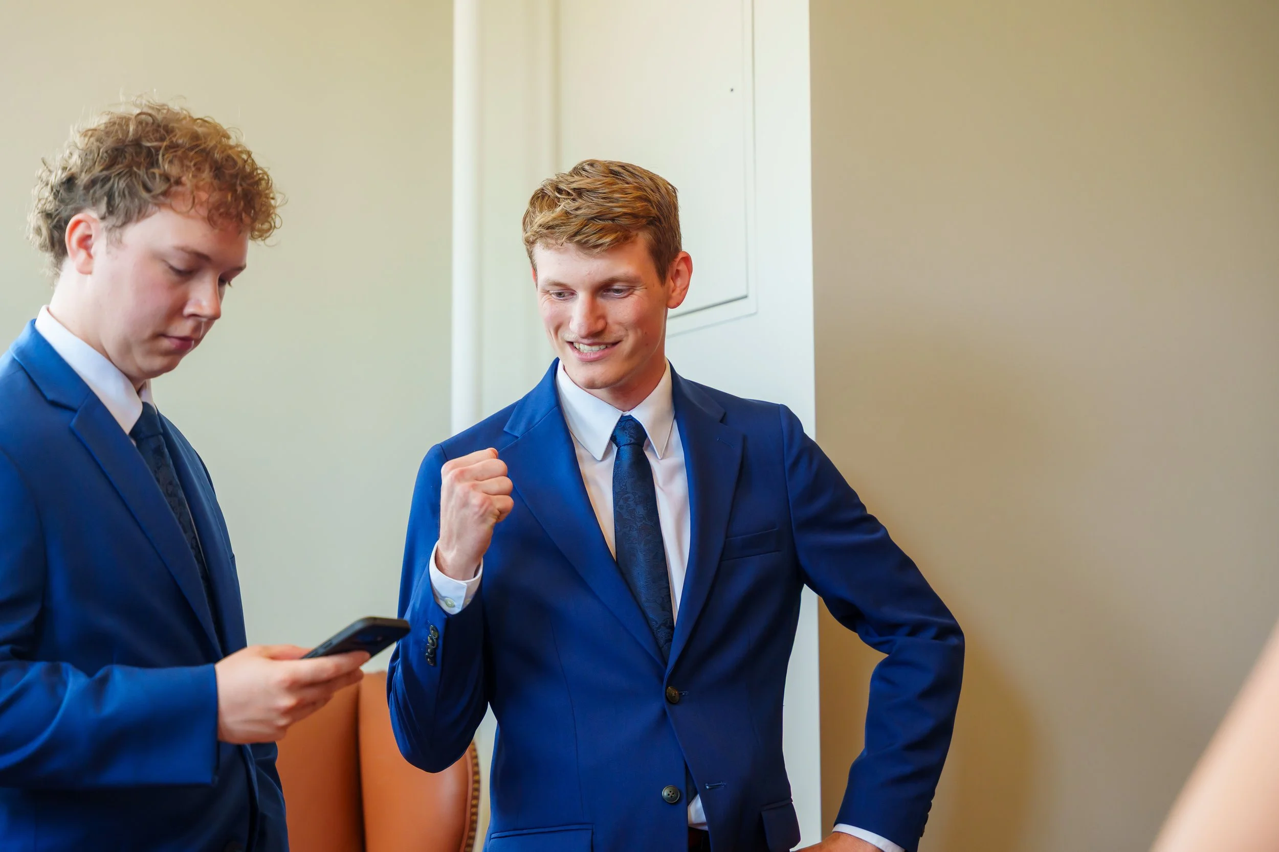 Two men in blue suits, one looking down at a phone and the other smiling and making a fist gesture, are standing in a room with beige walls.