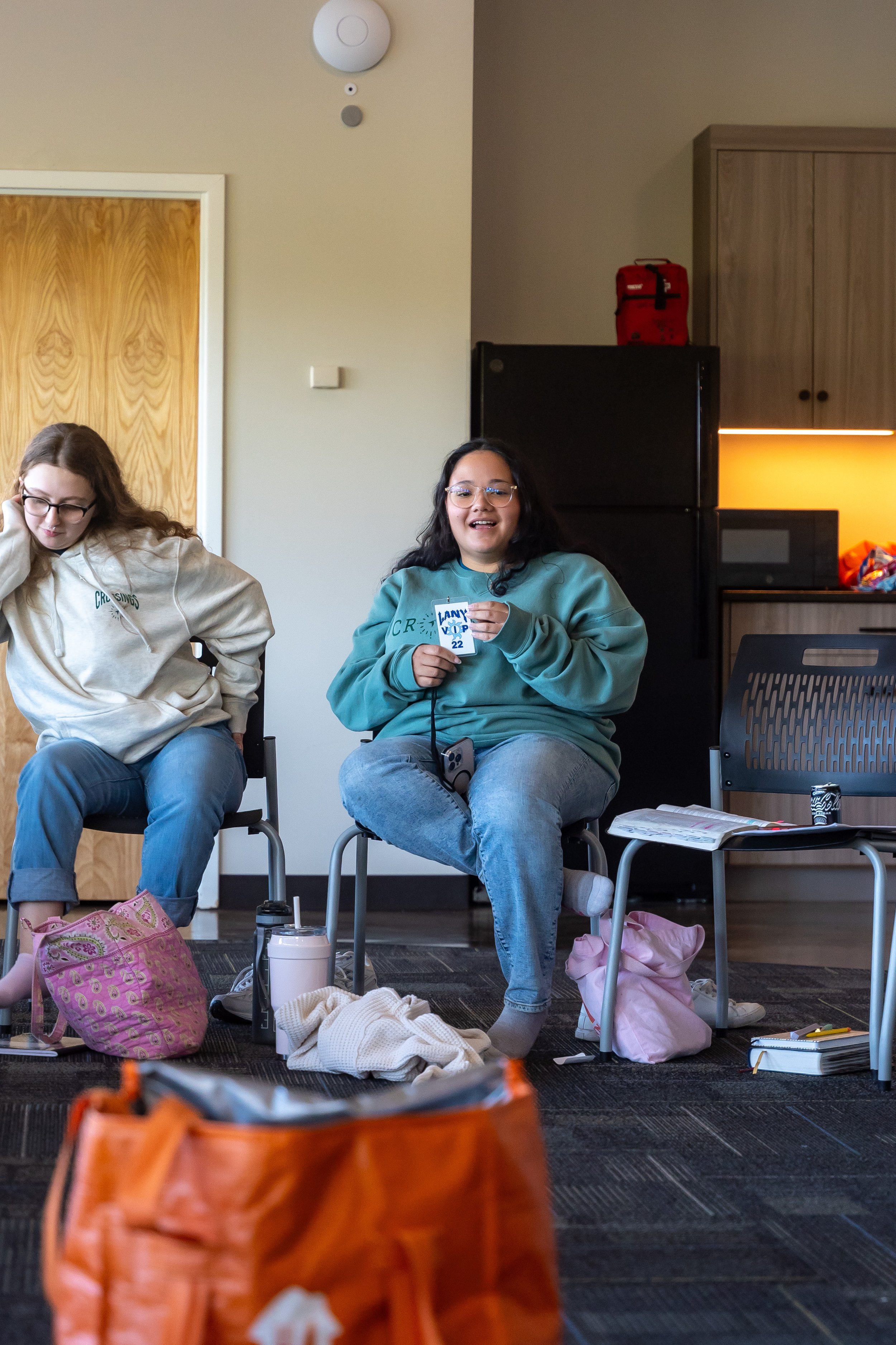 Two women sitting in chairs, surrounded by bags and personal items, in a room with a door, a black refrigerator, and a microwave. One woman with glasses and curly dark hair is smiling and holding a card, the other woman with glasses and long hair is 
