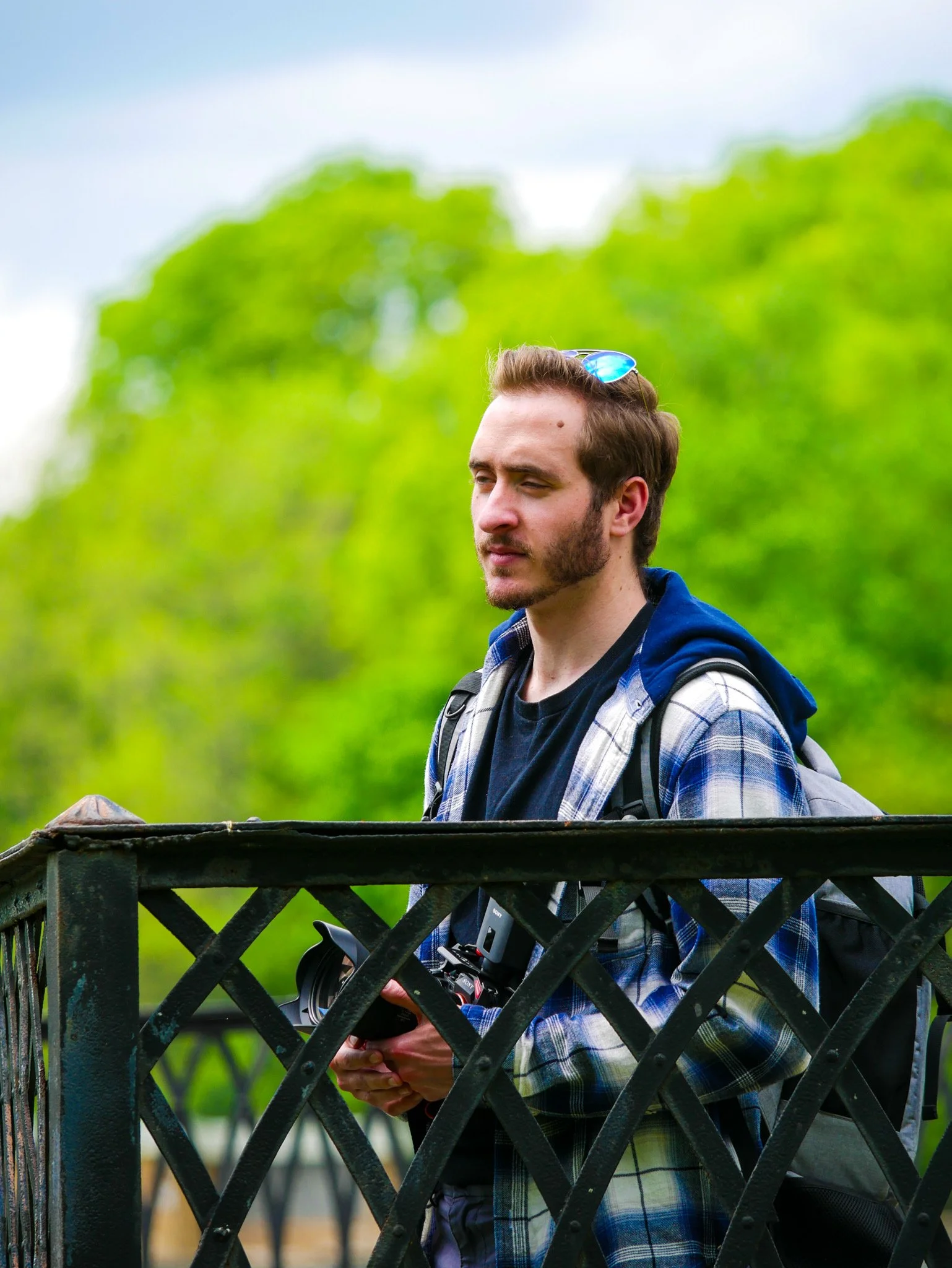 A man with short brown hair, beard, and sunglasses on his head, standing outdoors near a black wooden fence, holding a camera with a zoom lens, with green trees and a cloudy sky in the background.