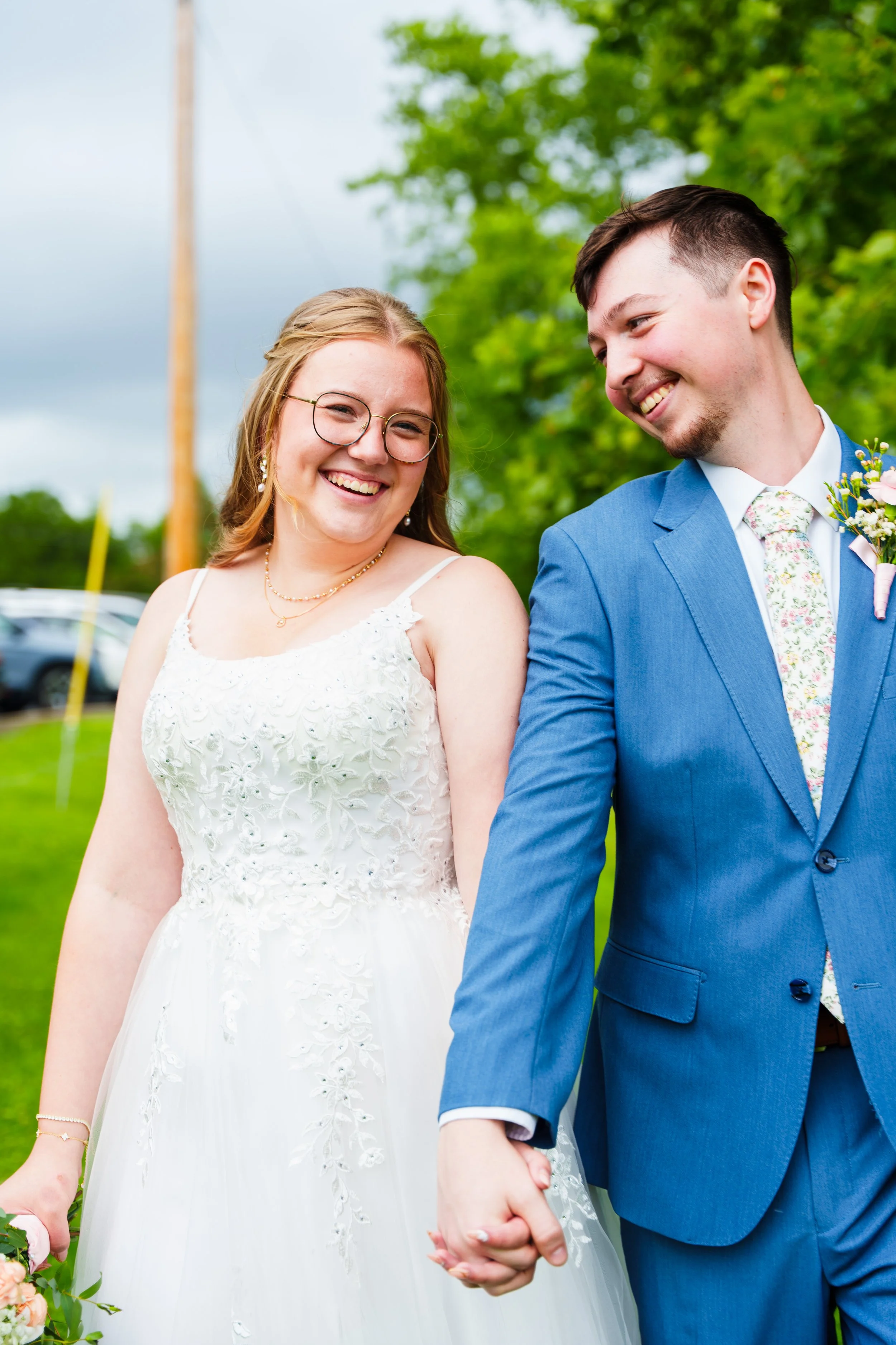 A smiling bride and groom holding hands outdoors with greenery in the background, celebrating their wedding.