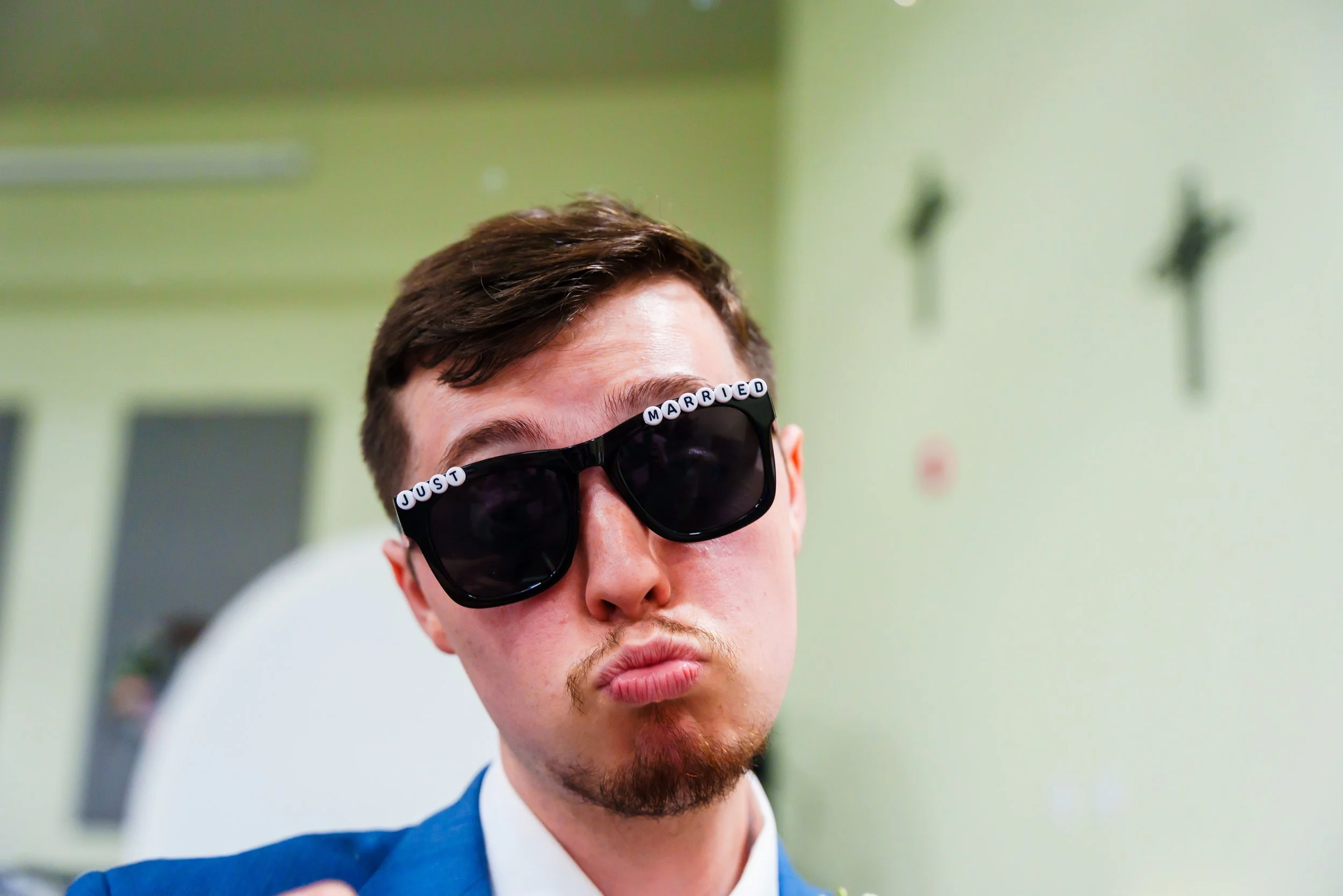 Close-up of a young man with brown hair and a beard wearing black sunglasses with white letter beads spelling 'JUST MARRIED' across the top. He is dressed in a blue suit and white shirt, standing indoors against a green wall.