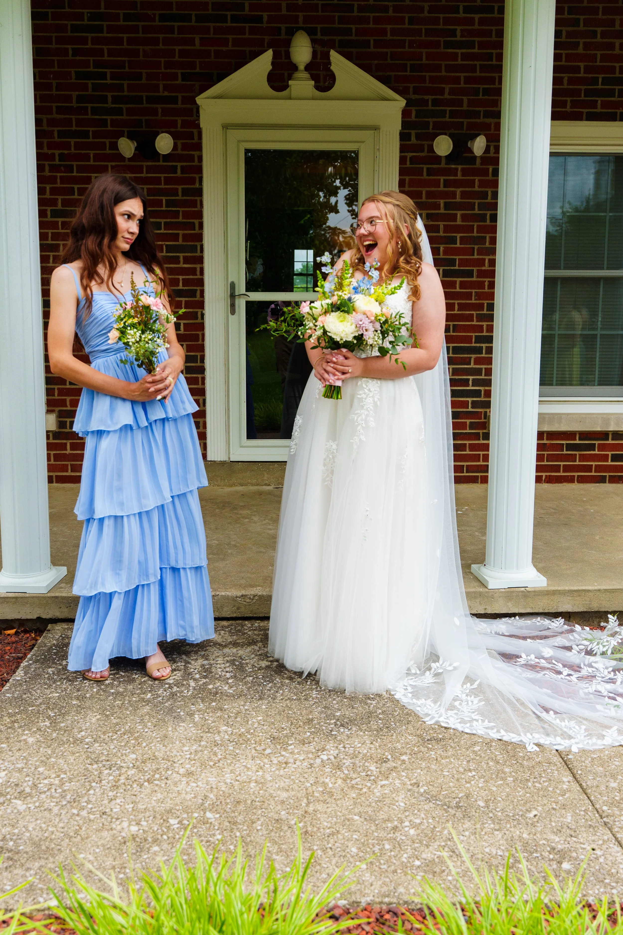 A bride in a white wedding dress and veil, holding a bouquet of white, pink, and purple flowers, laughing. Standing beside a bridesmaid in a blue dress, holding a smaller bouquet, making a skeptical or playful face. They are outdoors on a concrete po
