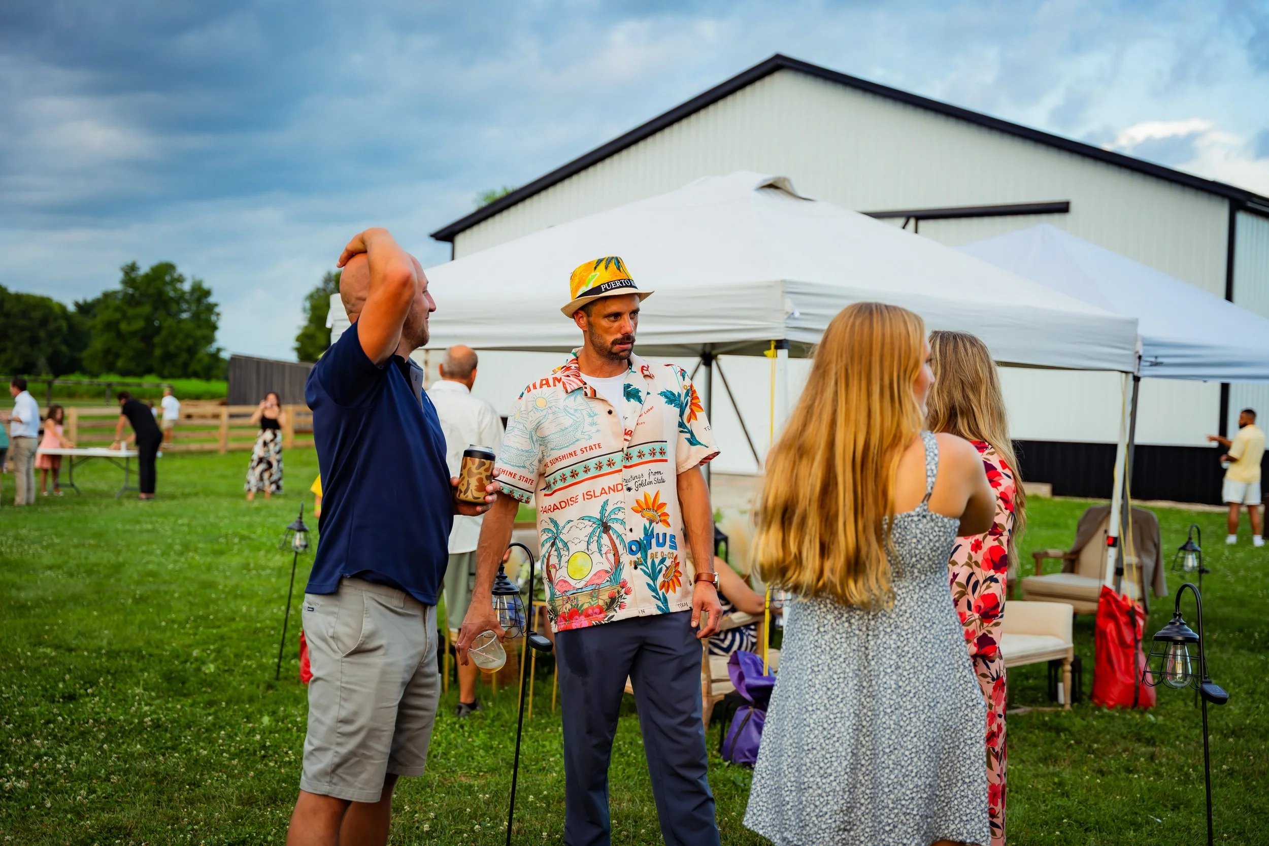 A group of people socializing outdoors on a grassy area with a white building and tents in the background. The sky is partly cloudy, and some individuals are dressed casually, with one man wearing a colorful tropical shirt and a hat.