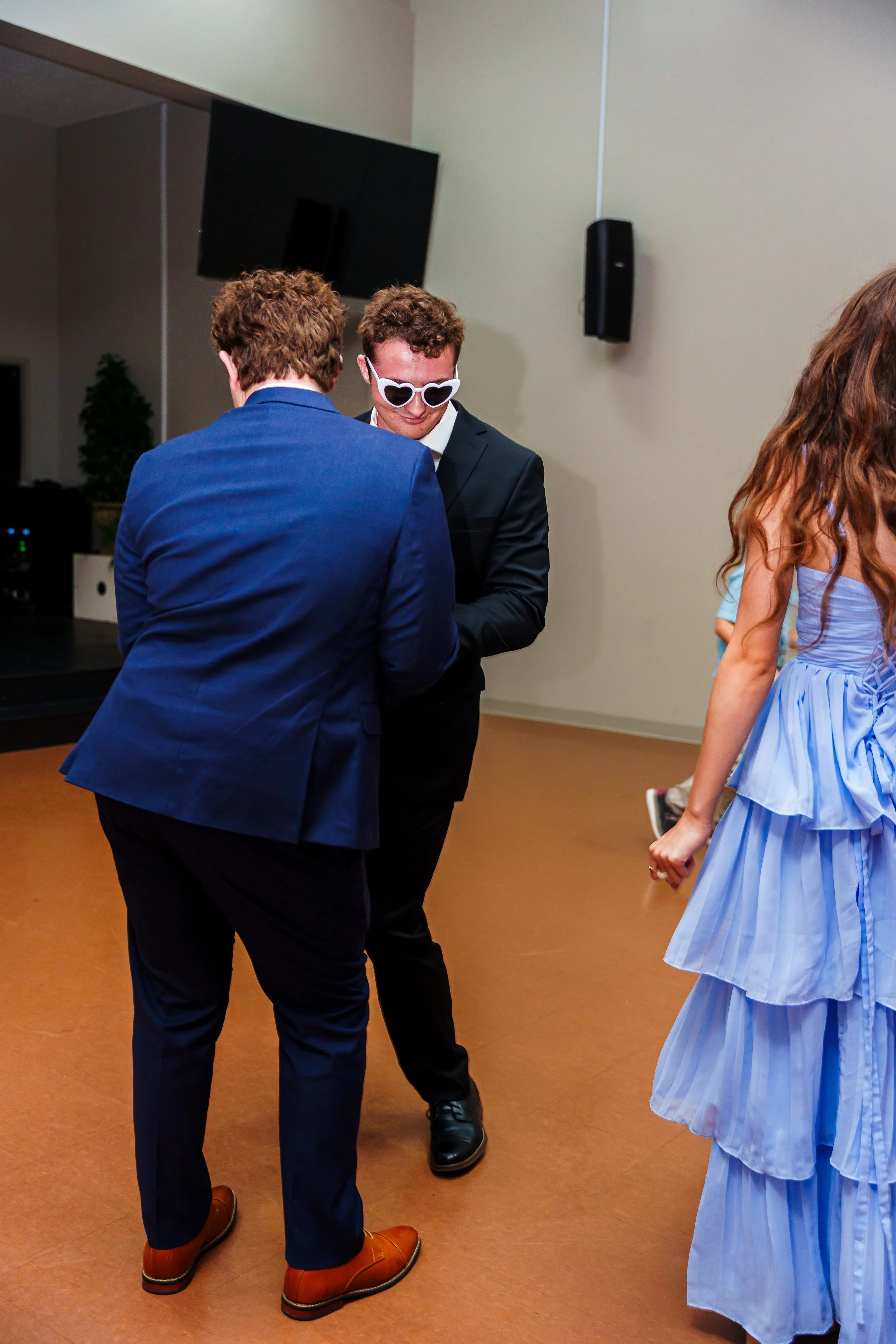 Group of people dancing at an indoor event, including a man in a black suit with white heart-shaped sunglasses, a woman in a light blue dress, and others in formal attire.