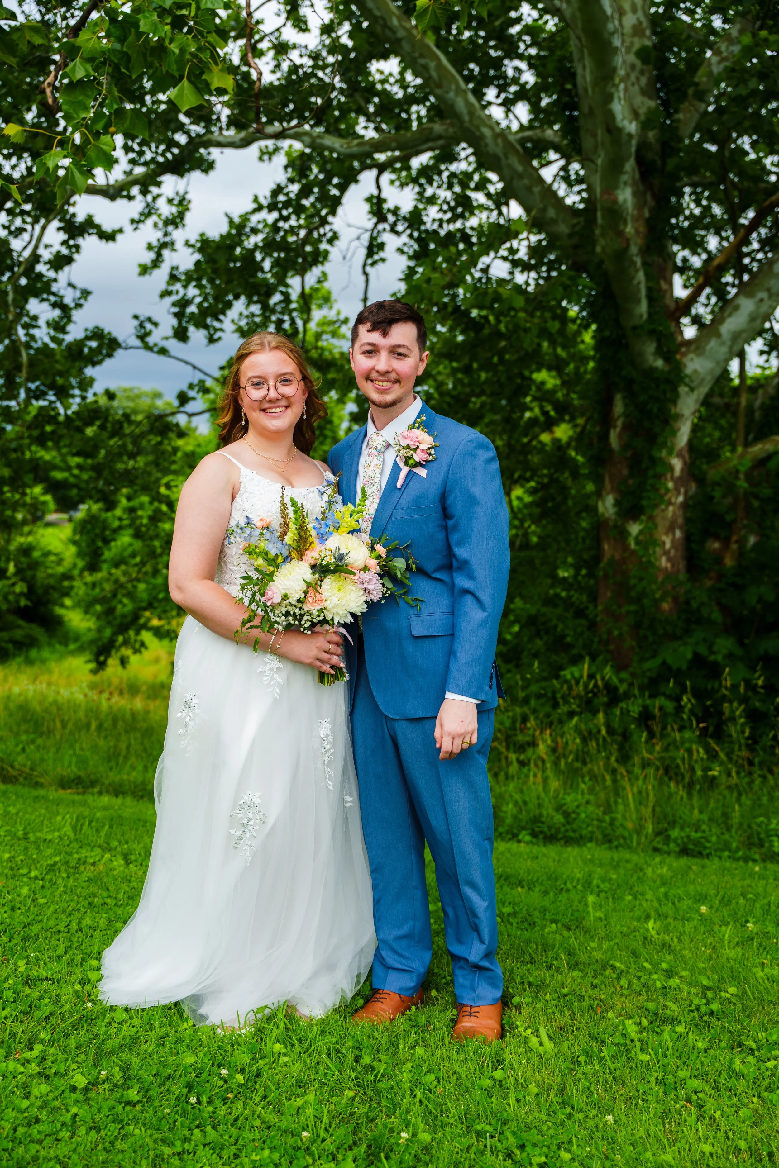A bride and groom standing outdoors on a grassy area with green trees in the background. The bride is wearing a white wedding dress and glasses, holding a bouquet of flowers. The groom is dressed in a blue suit and tie, smiling with his arm around th