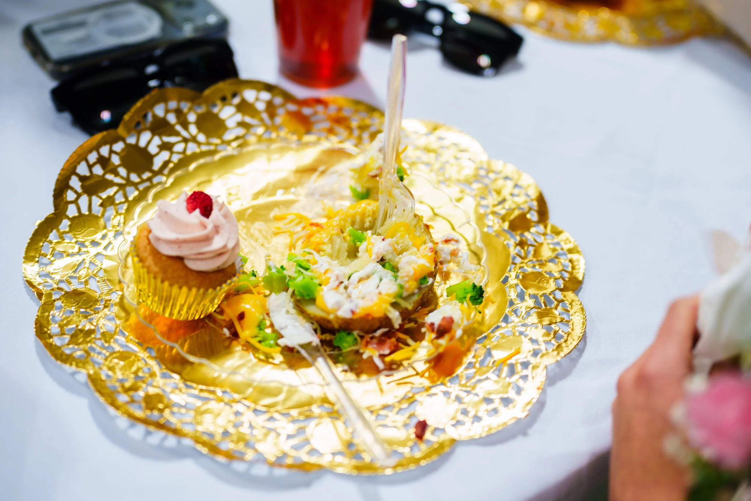 A birthday cake with yellow frosting and colorful decorations on a gold doily, with a cupcake topped with pink frosting and a cherry, a red drink, and a pair of glasses on a white table.