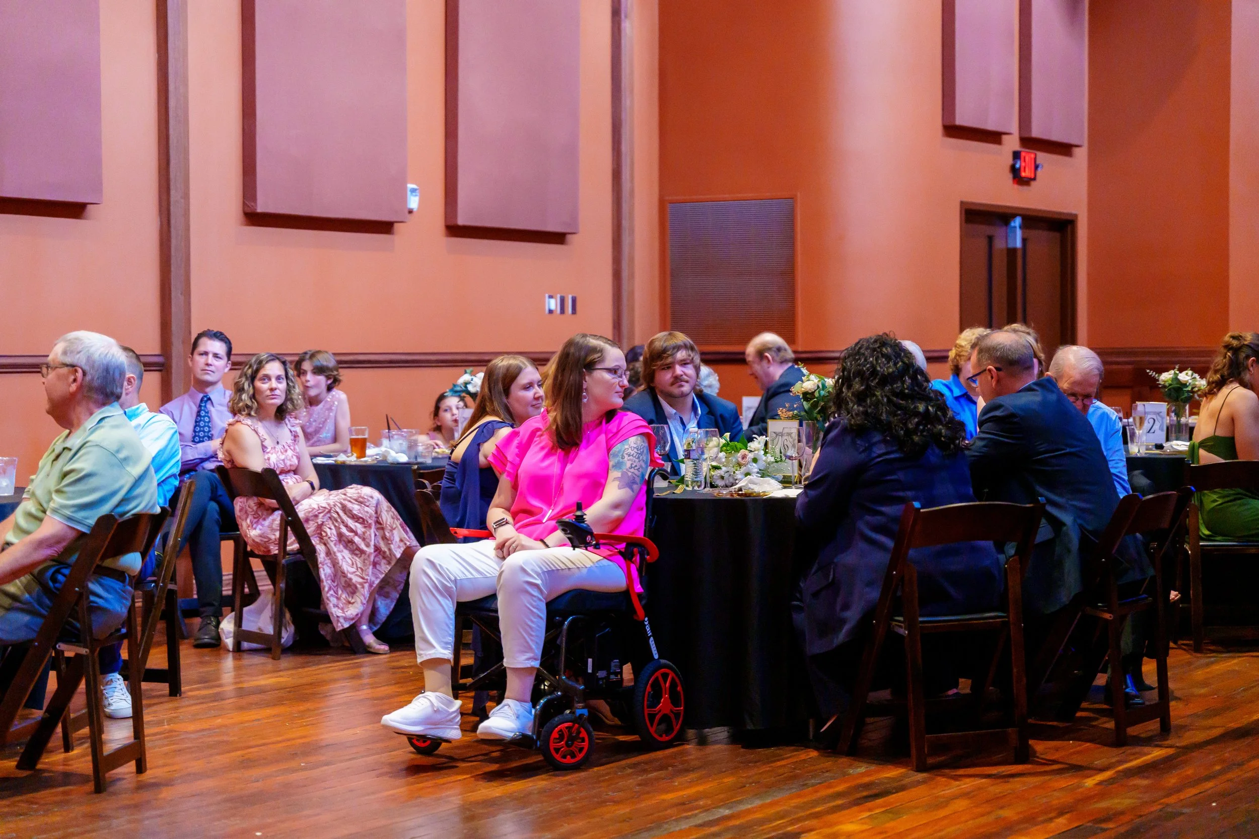 Guests seated at tables in a banquet hall attending a formal event or celebration, with orange walls and wooden floors.