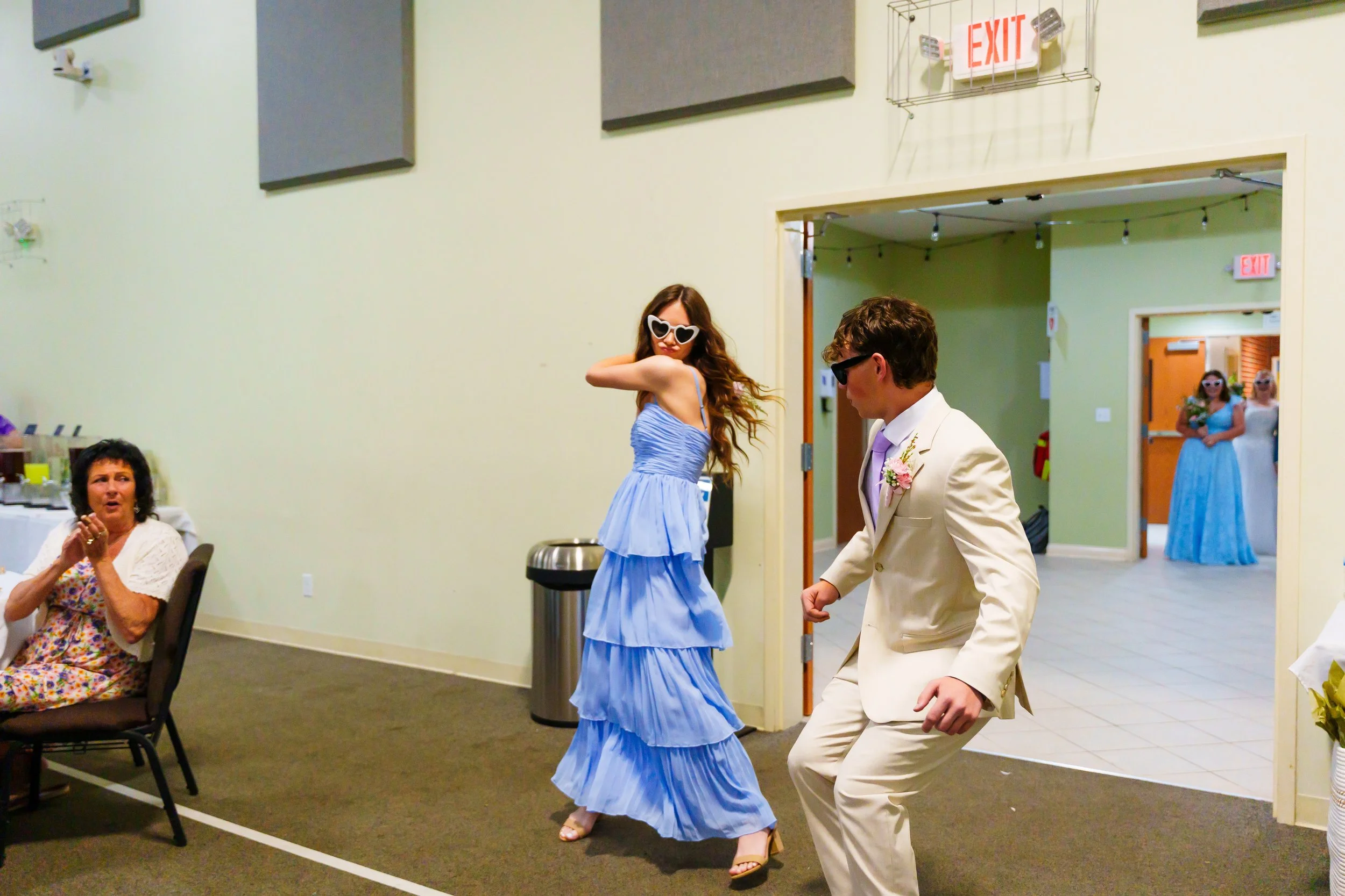 A young woman in a blue dress and heart-shaped sunglasses dancing at a wedding reception. An older woman is sitting at a table nearby, and a man in a cream suit and black sunglasses is also dancing. In the background, there is a doorway, and two wome