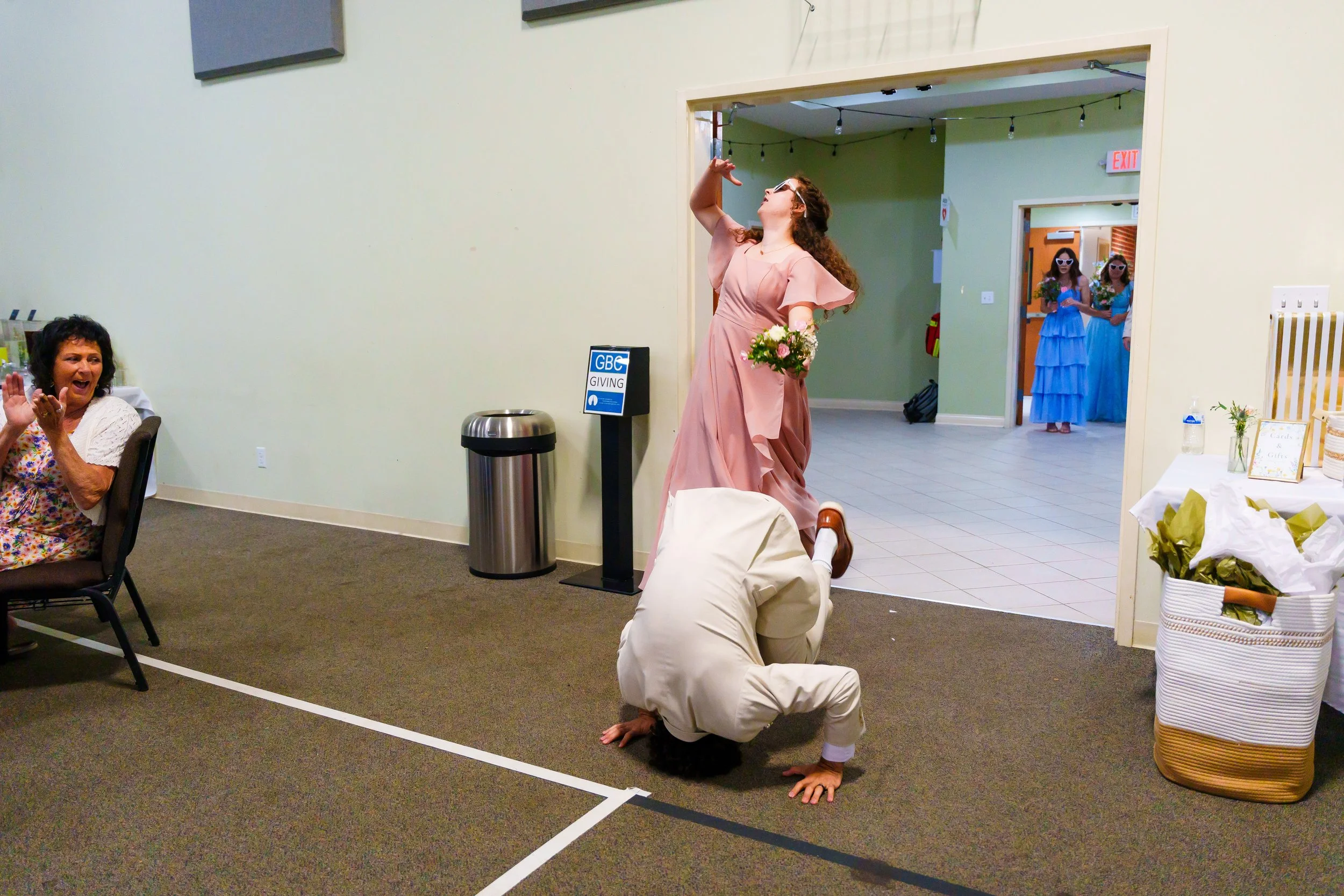 A person in a beige suit is performing a handstand in a room decorated for a celebration, with a woman sitting at a table to the left and two women in blue dresses holding flowers in the background.