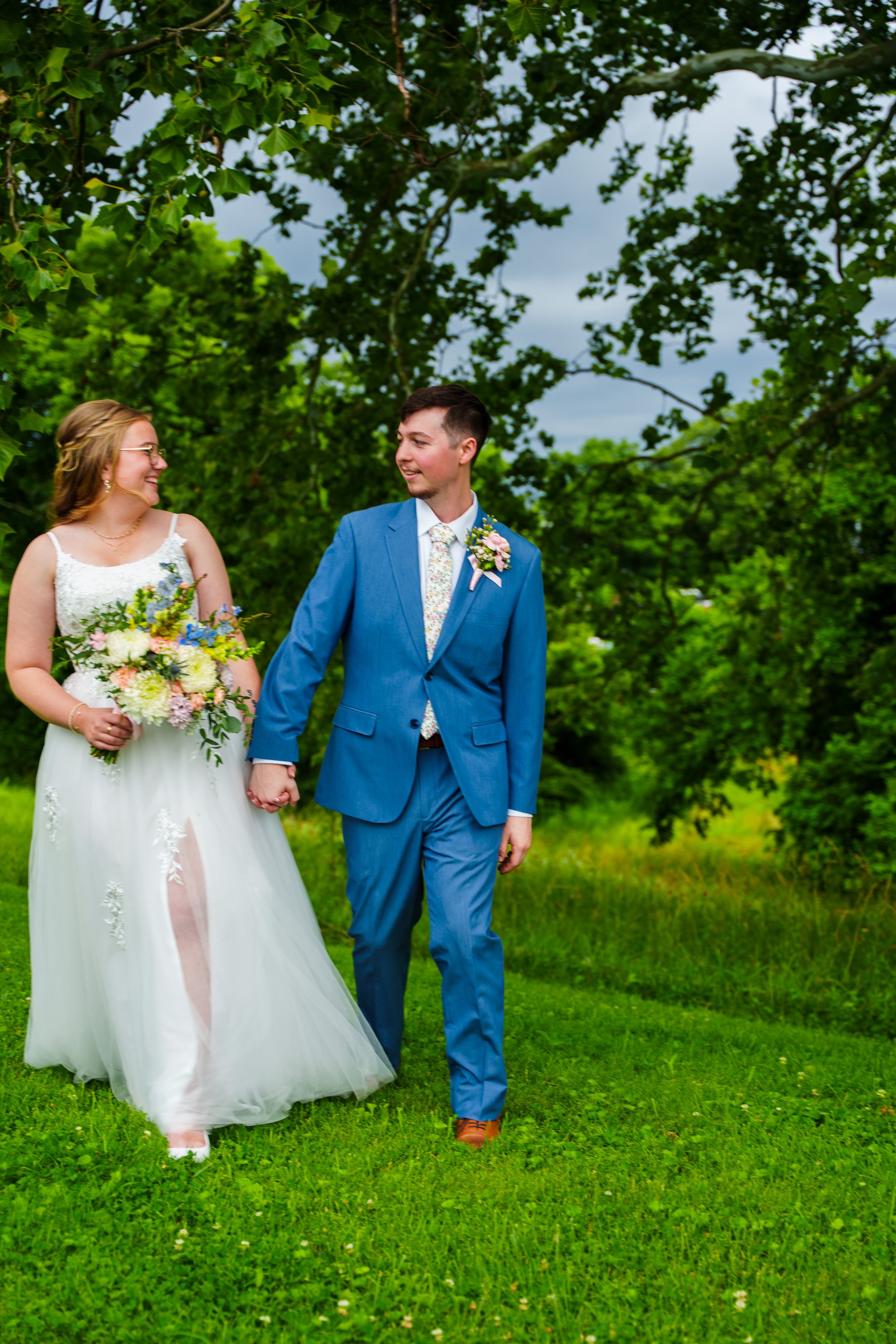 A bride and groom holding hands and smiling at each other outdoors on a grassy field with trees and cloudy sky in the background.