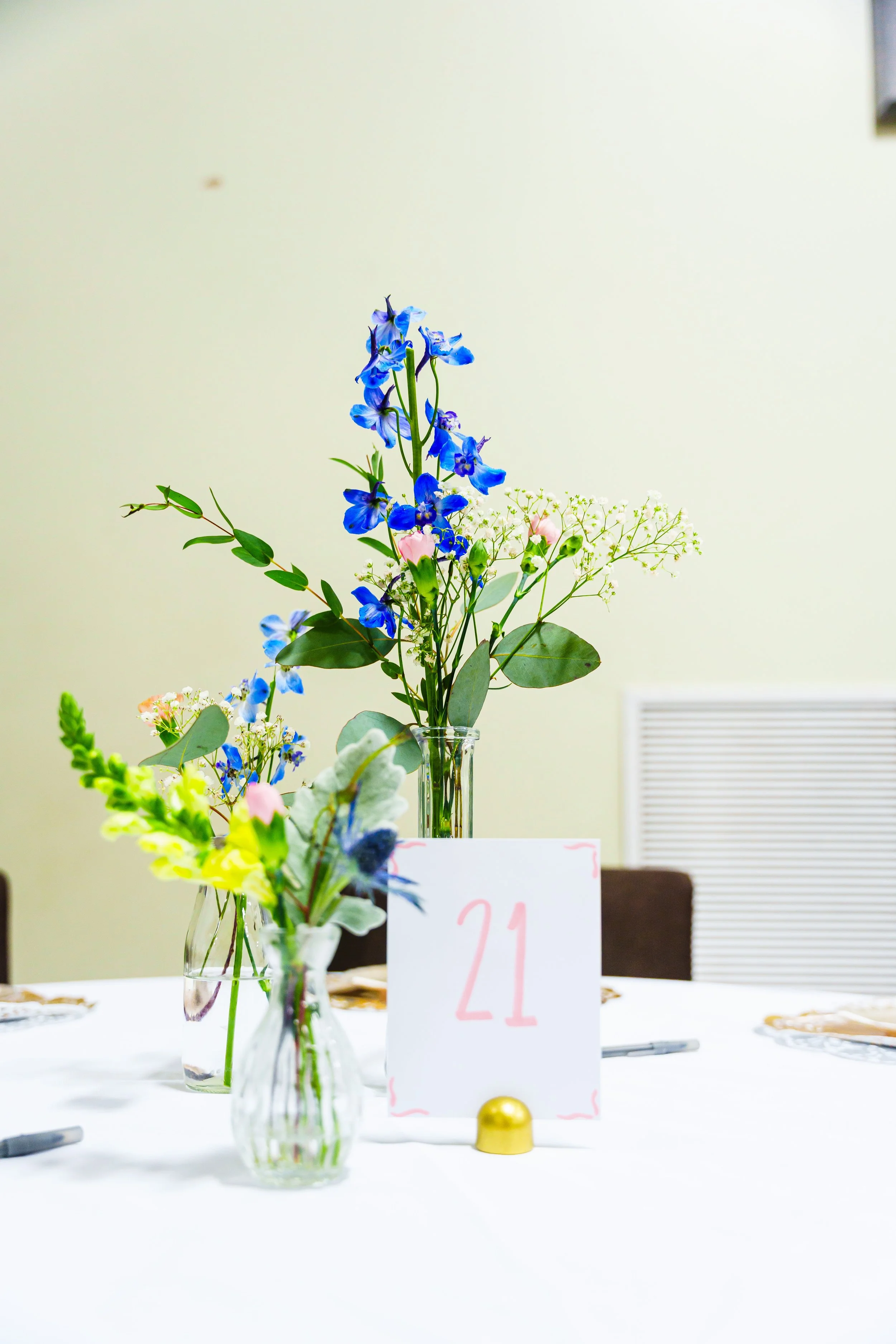 A floral arrangement with blue, pink, and white flowers in a glass vase on a white table. A table number card with the number 21 is in front of the arrangement, and a small gold decorative object is at the base of the card. The background is plain wi