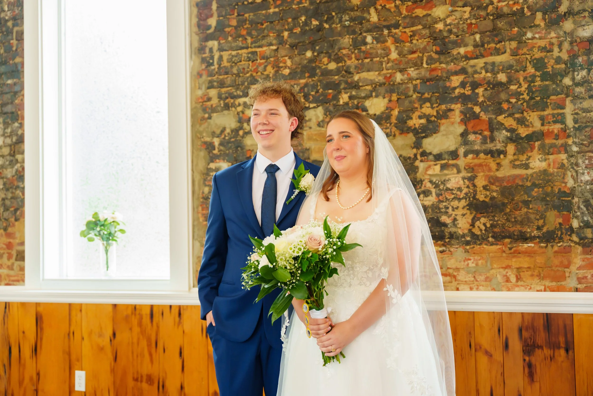 A bride and groom standing together during their wedding ceremony. The groom is wearing a blue suit with a white shirt and a dark blue tie, and has a white floral boutonniere. The bride is dressed in a white lace wedding gown, with a pearl necklace, 