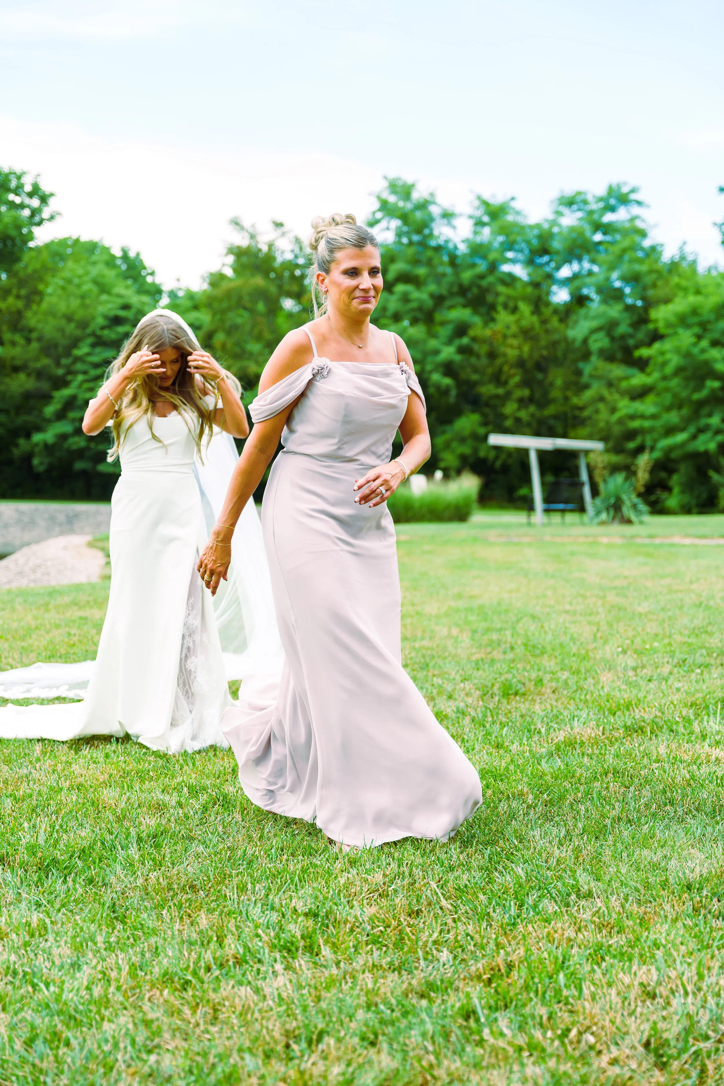 A woman in a light-colored dress walking across a grassy field with trees in the background, accompanied by a girl in a white dress.