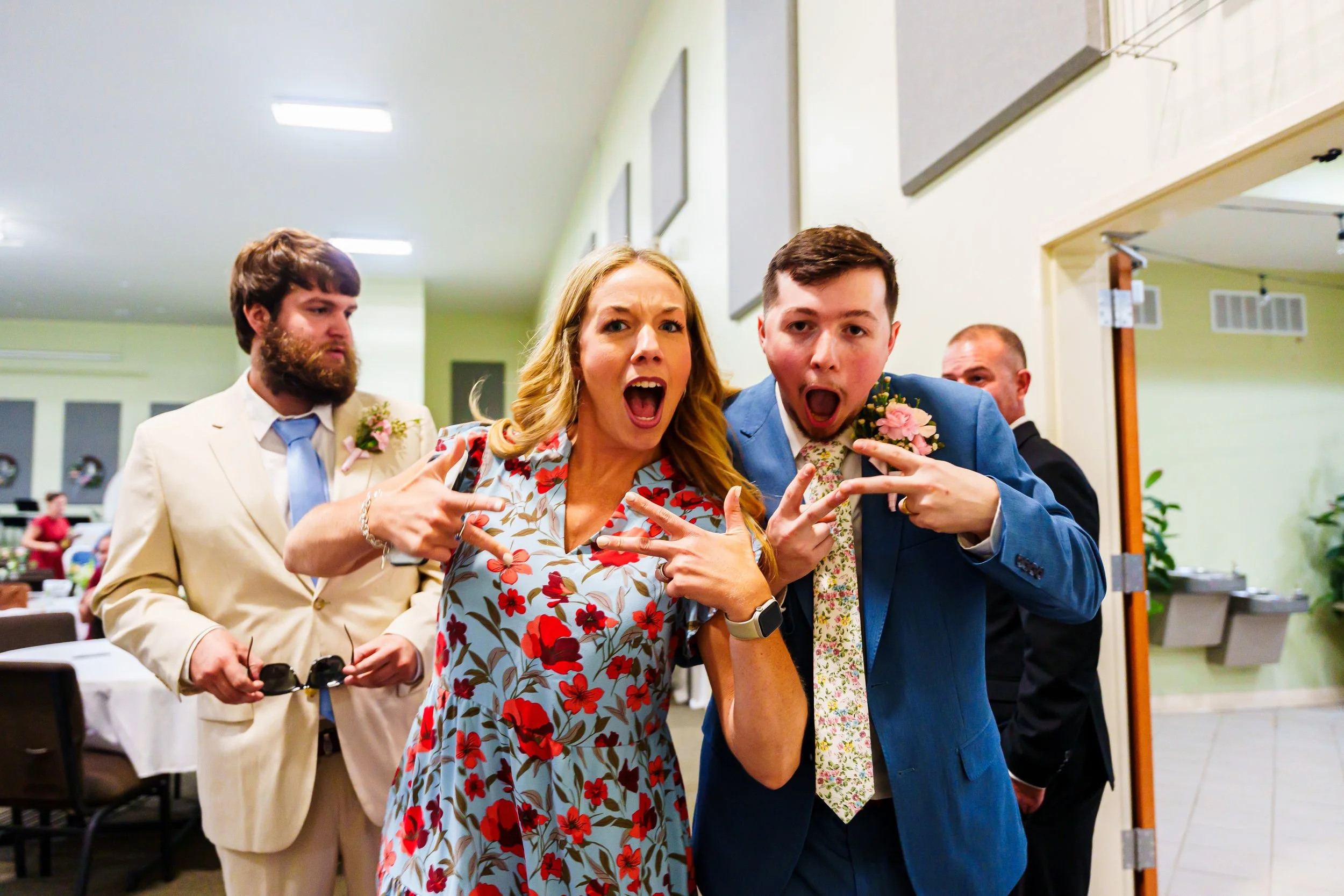 A group of people at what appears to be a wedding reception. Two men and a woman in the foreground are posing playfully for the camera, making peace signs and wide-open mouths. The woman is wearing a floral dress, and one man is in a blue suit. The b