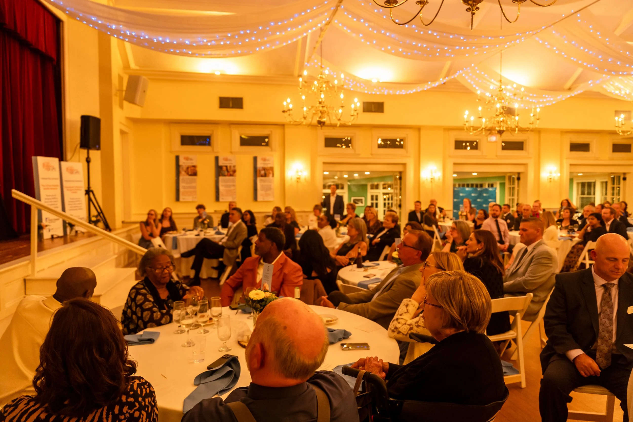 A large indoor banquet hall filled with round tables of people dressed in formal attire, with decorative lighting and chandeliers hanging from the ceiling, and informational posters on the walls.