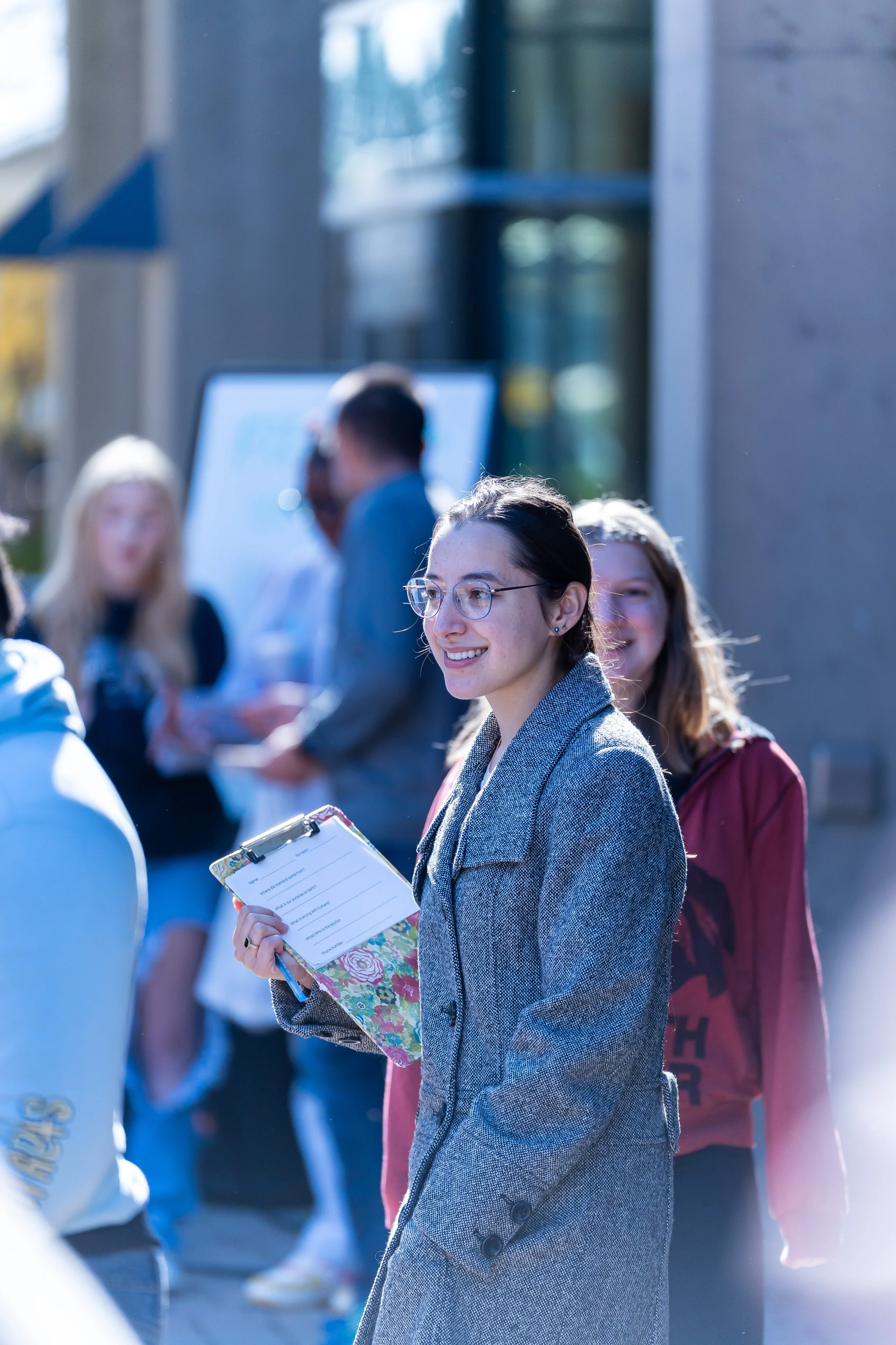 A young woman with glasses, dark hair, and a grey blazer, holding a clipboard, smiling and standing outdoors among people.