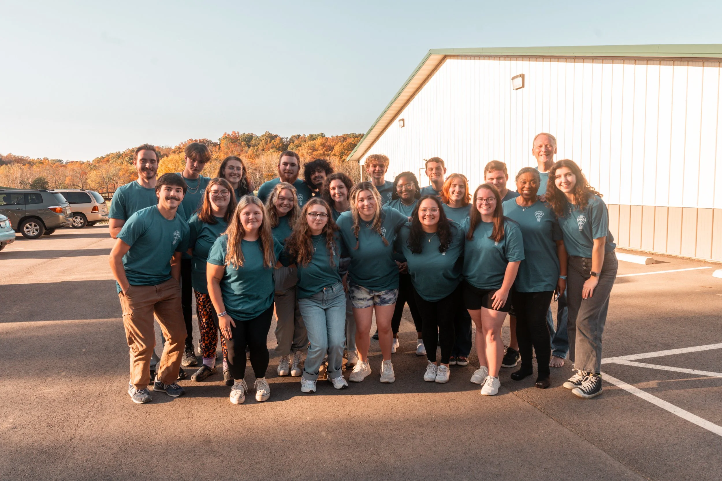 Group of people standing outdoors, smiling for a photo, in front of a white building and parked cars, with trees in the background during fall.
