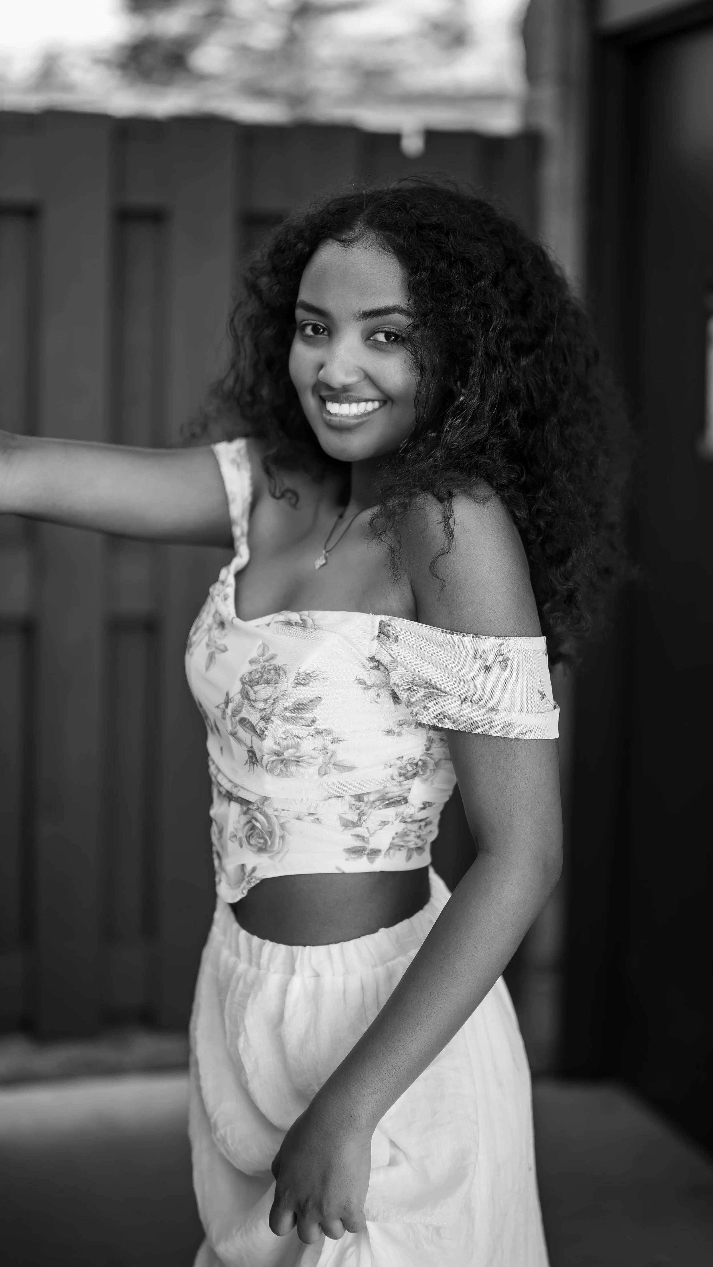 A young woman with curly hair smiling and looking at the camera in a black and white photo, wearing an off-the-shoulder floral top and light-colored pants.