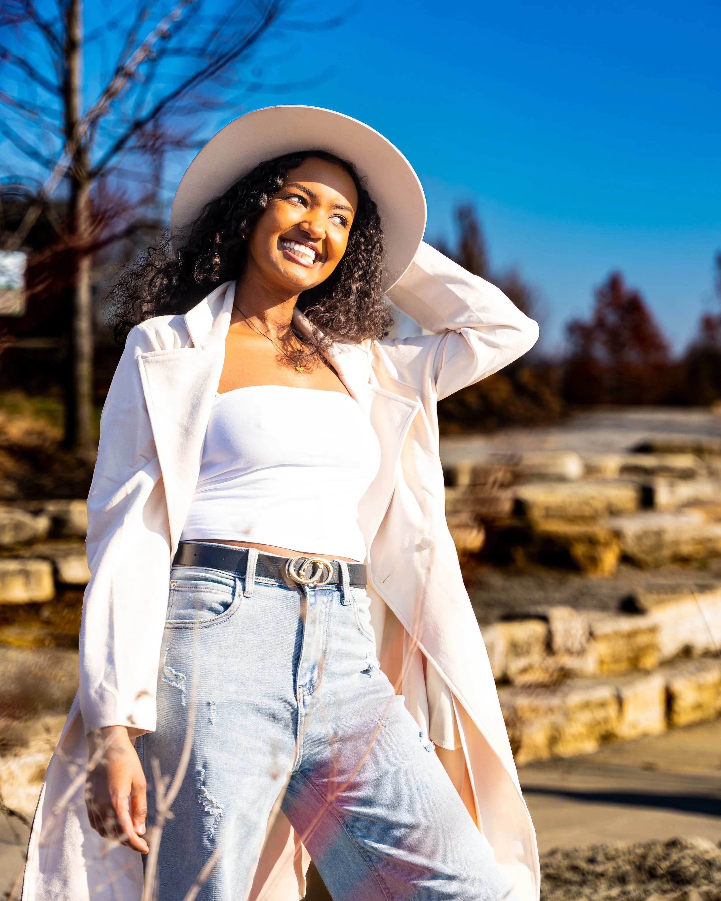 A woman smiling outdoors under a clear blue sky, wearing a white hat, white top, light jeans with a Gucci belt, and a light-colored blazer draped over her shoulders, with trees and stone steps in the background.
